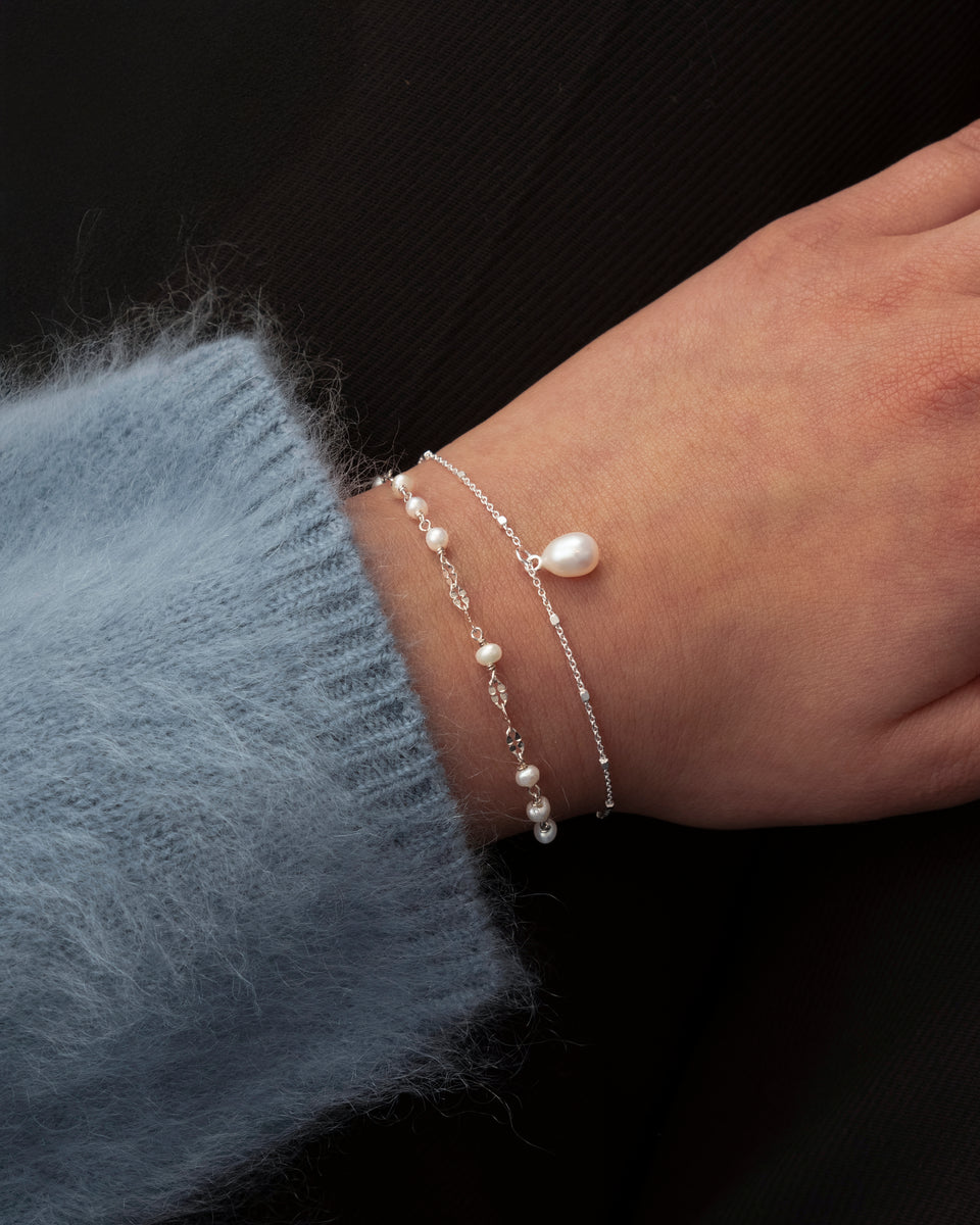 A close-up of a model's wrist wearing the Treasure Single Pearl Bracelet in silver, which features a delicate chain and a single teardrop-shaped pearl, layered with the silver Aurelia Bracelet adorned with multiple smaller pearls.