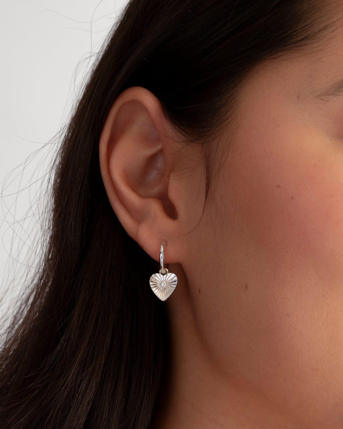 A close-up shot of a model wearing the Aurelia Earring in silver, featuring a small hoop with a dangling, textured heart-shaped pendant that holds a small freshwater pearl at its center.