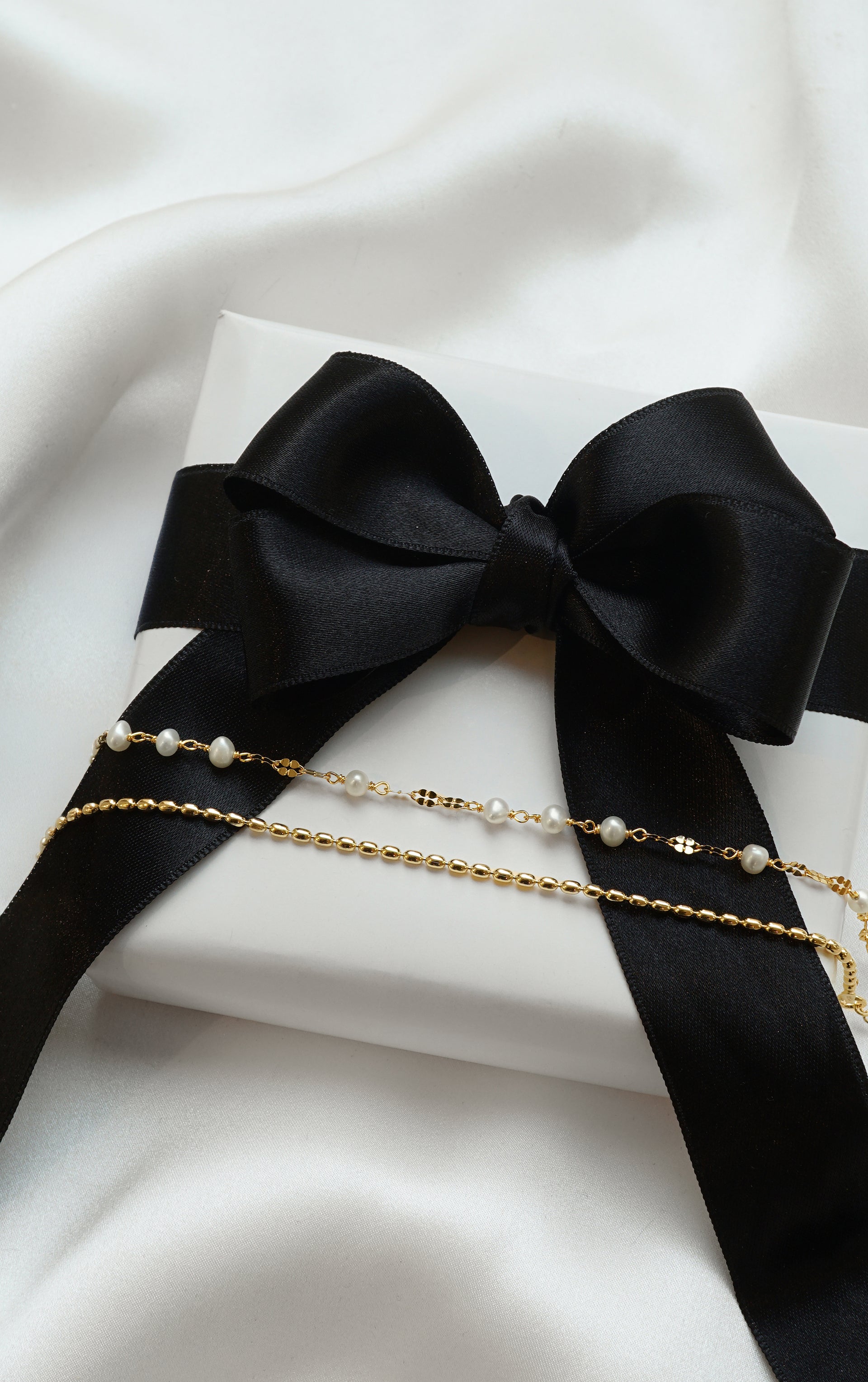 A product shot of the Aurelia & Lunetta Bracelet Set in gold, resting on a white gift box wrapped with a black ribbon. The set features one bracelet with a delicate chain and freshwater pearls, and another with a beaded gold chain design.