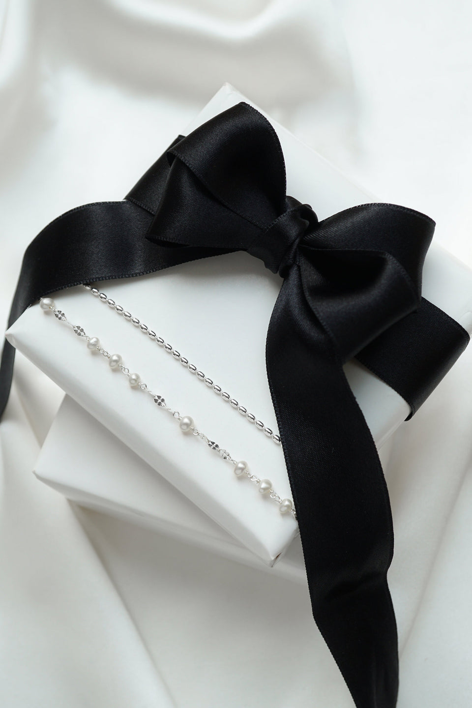 A product shot of the Aurelia & Lunetta Bracelet Set in silver, displayed on a white gift box tied with a black ribbon, showcasing both the freshwater pearl Aurelia bracelet and the sleek Lunetta chain bracelet.