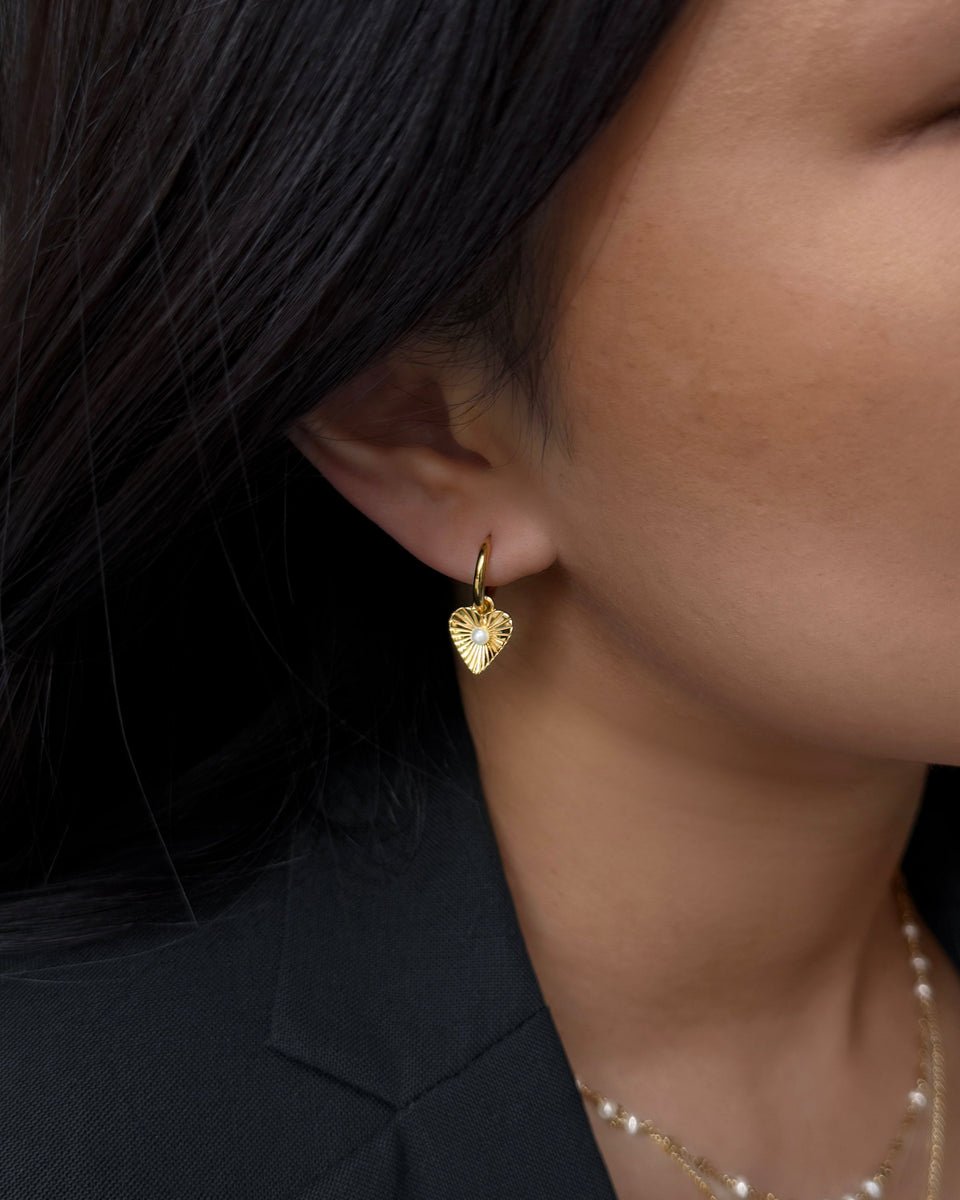 A close-up shot of a model wearing the Aurelia Earring in gold, featuring a small hoop with a textured, heart-shaped pendant and a central freshwater pearl. She is also wearing a layered gold necklace with pearls.