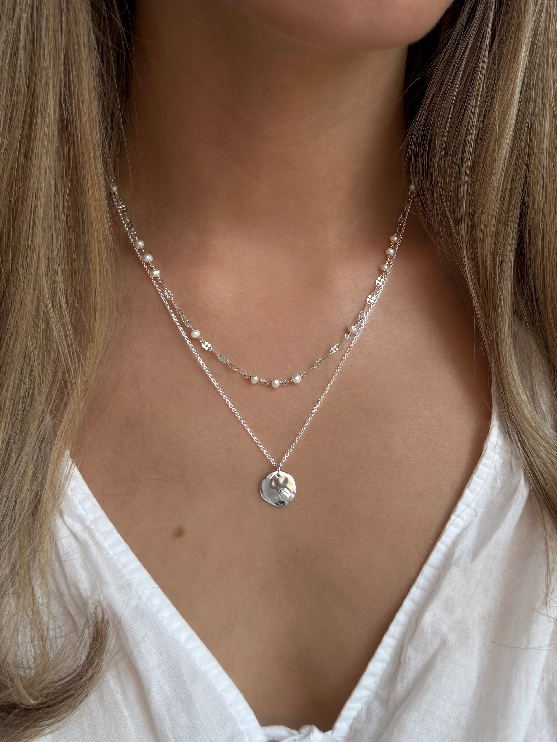 A close-up on the neck of a model wearing two layered silver necklaces. The longer is the Paxton Necklace with a hammered round pendant and a baroque pearl, styled with the shorter Aurelia pearl necklace.