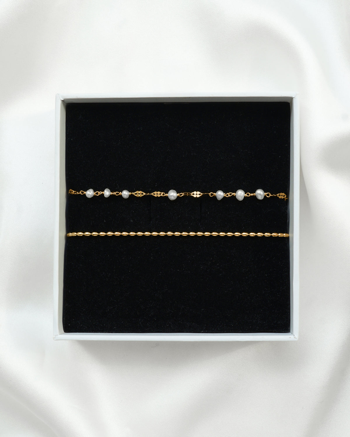 A product shot of the Aurelia & Lunetta Bracelet Set in gold, displayed in a white gift box. The set includes a delicate chain bracelet with freshwater pearls and a twisted gold chain bracelet.