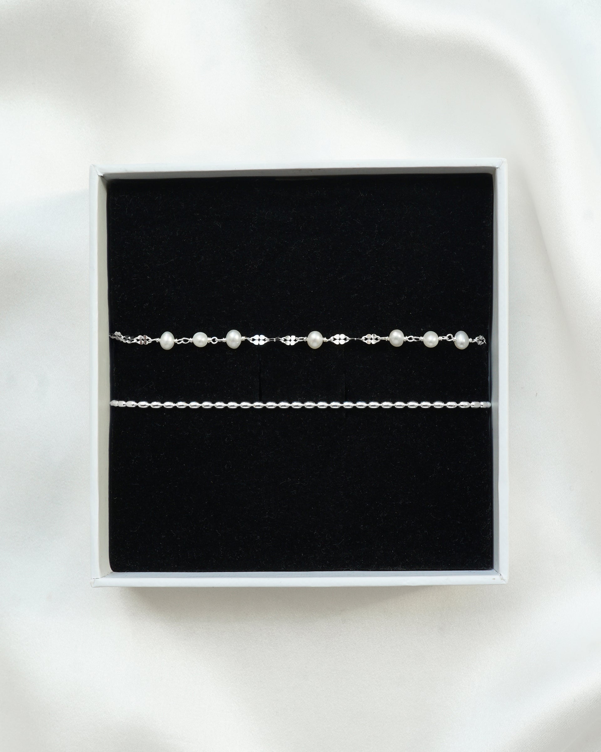 A product shot of the Aurelia & Lunetta Bracelet Set in silver, presented in a white gift box. The set includes the Aurelia bracelet, featuring a fine chain with freshwater pearls, and the Lunetta bracelet, a simple silver chain.