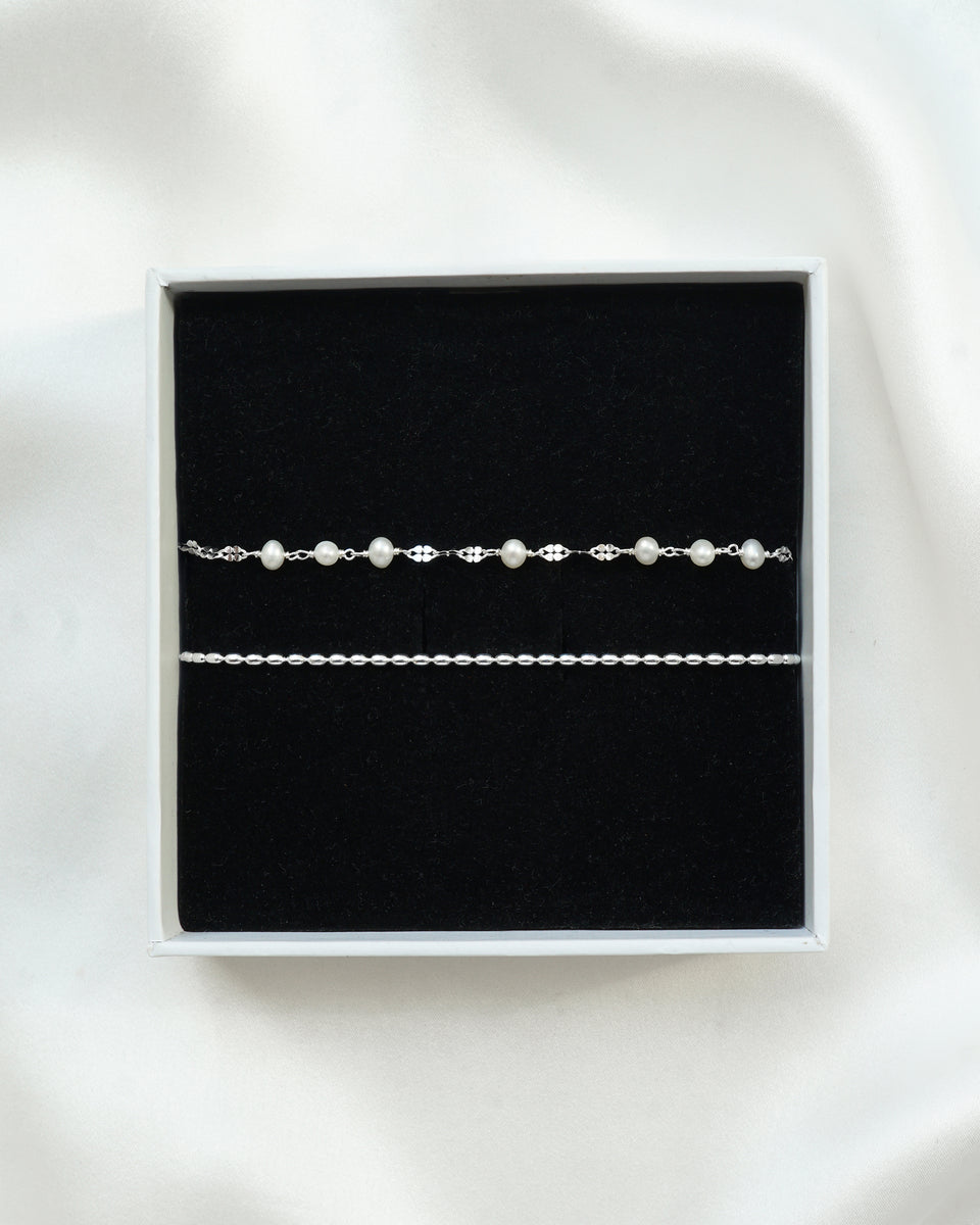 A product shot of the Aurelia & Lunetta Bracelet Set in silver, presented in a white gift box. The set includes the Aurelia bracelet, featuring a fine chain with freshwater pearls, and the Lunetta bracelet, a simple silver chain.