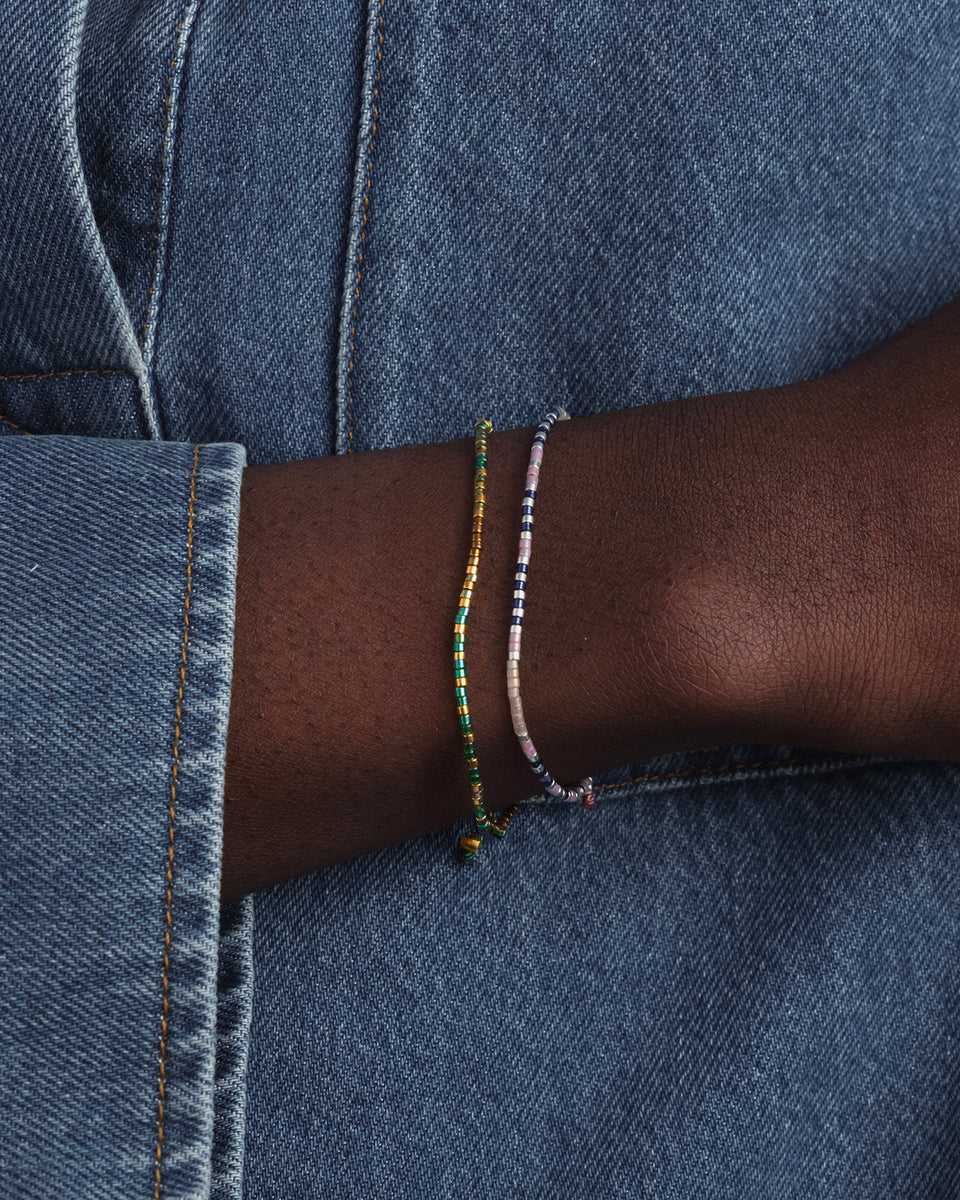 A close-up of a model's wrist wearing two stacked Syster P Code Bracelets: the 'Just Breathe' bracelet with pink, blue, and silver beads, and the 'Good Vibes' bracelet with gold and green beads.