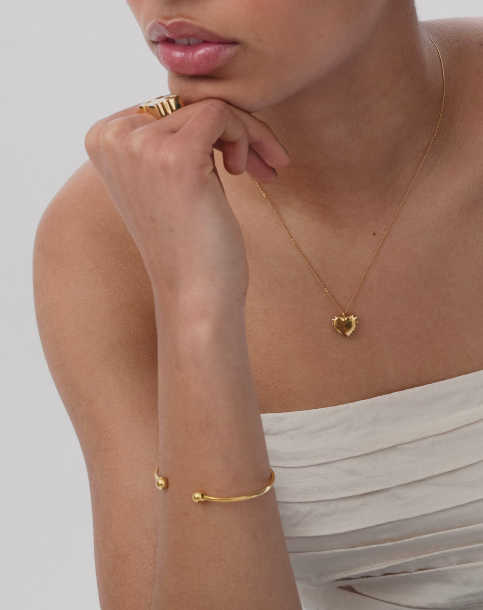 A model wearing the gold Strict Ball Bangle on her wrist, styled with a gold necklace featuring a spiked heart pendant and a chunky gold ring.