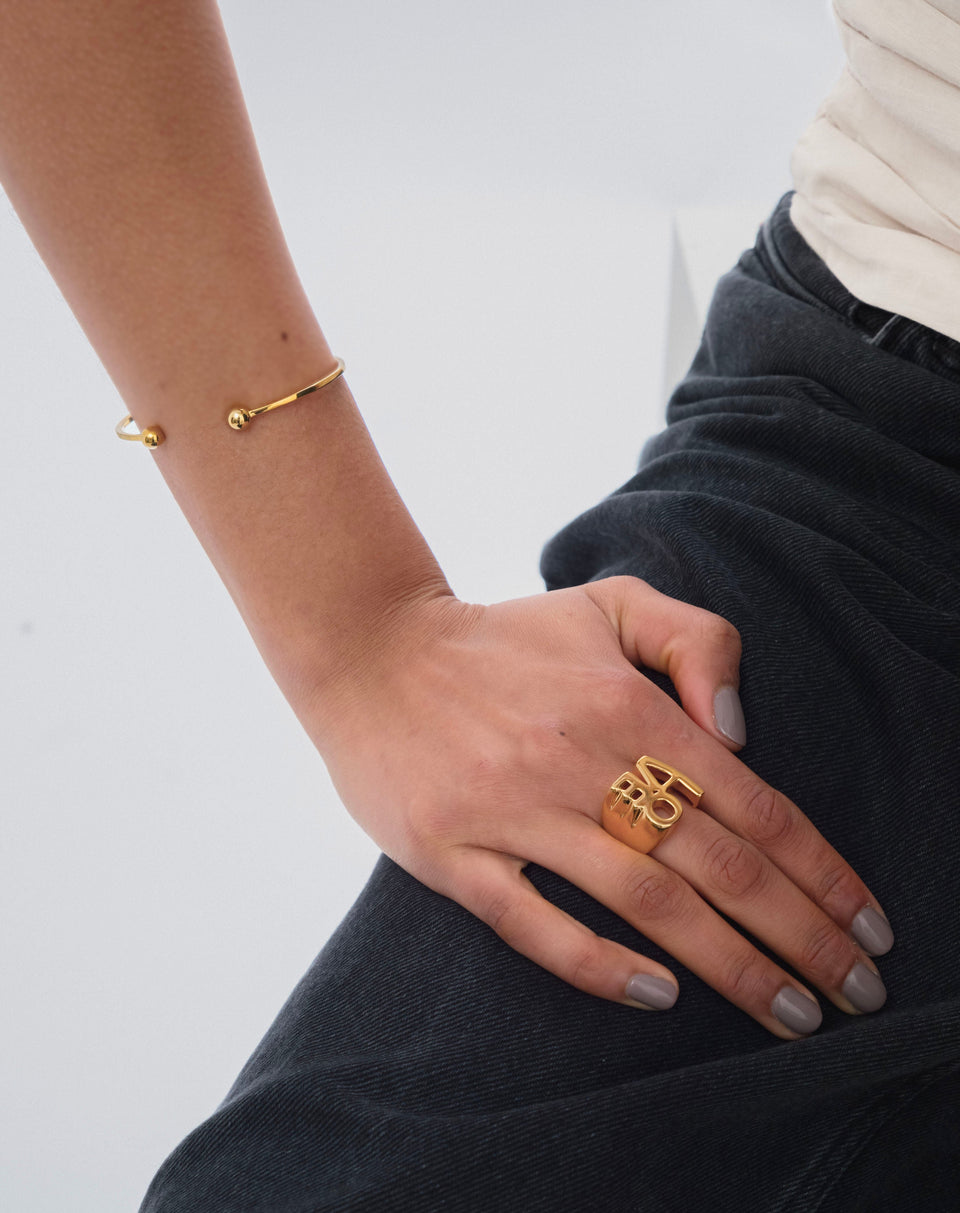 A close-up of a model wearing the Strict Ball Bangle Gold, a thin cuff with balls at the ends, paired with a large gold statement ring.