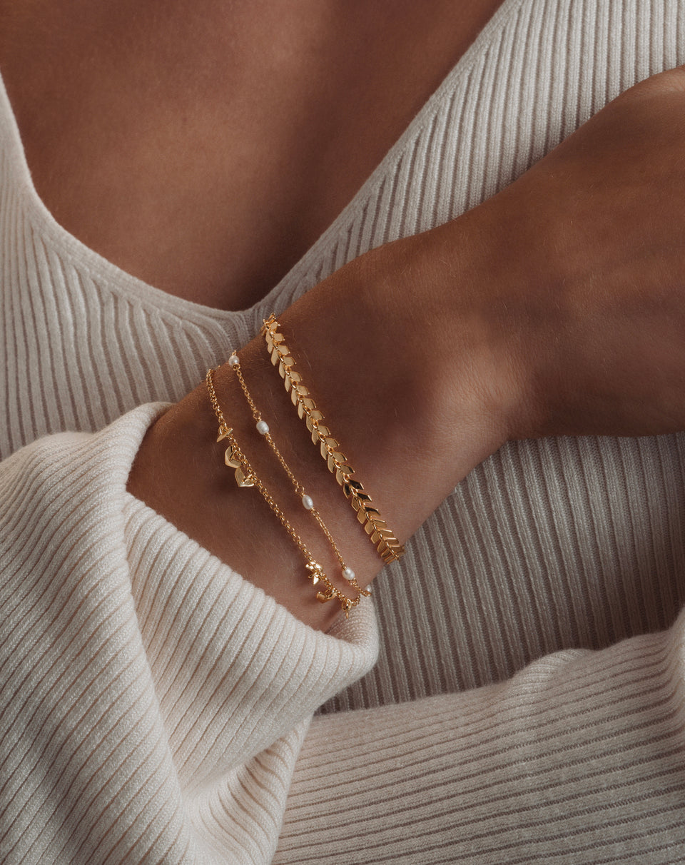 A model's wrist, wearing a stack of three gold bracelets against the sleeve of a cream ribbed top. The stack includes the prominent Layers Olivia Bracelet with its leaf-like design, a delicate chain with small pearls, and a third fine chain with charms.
