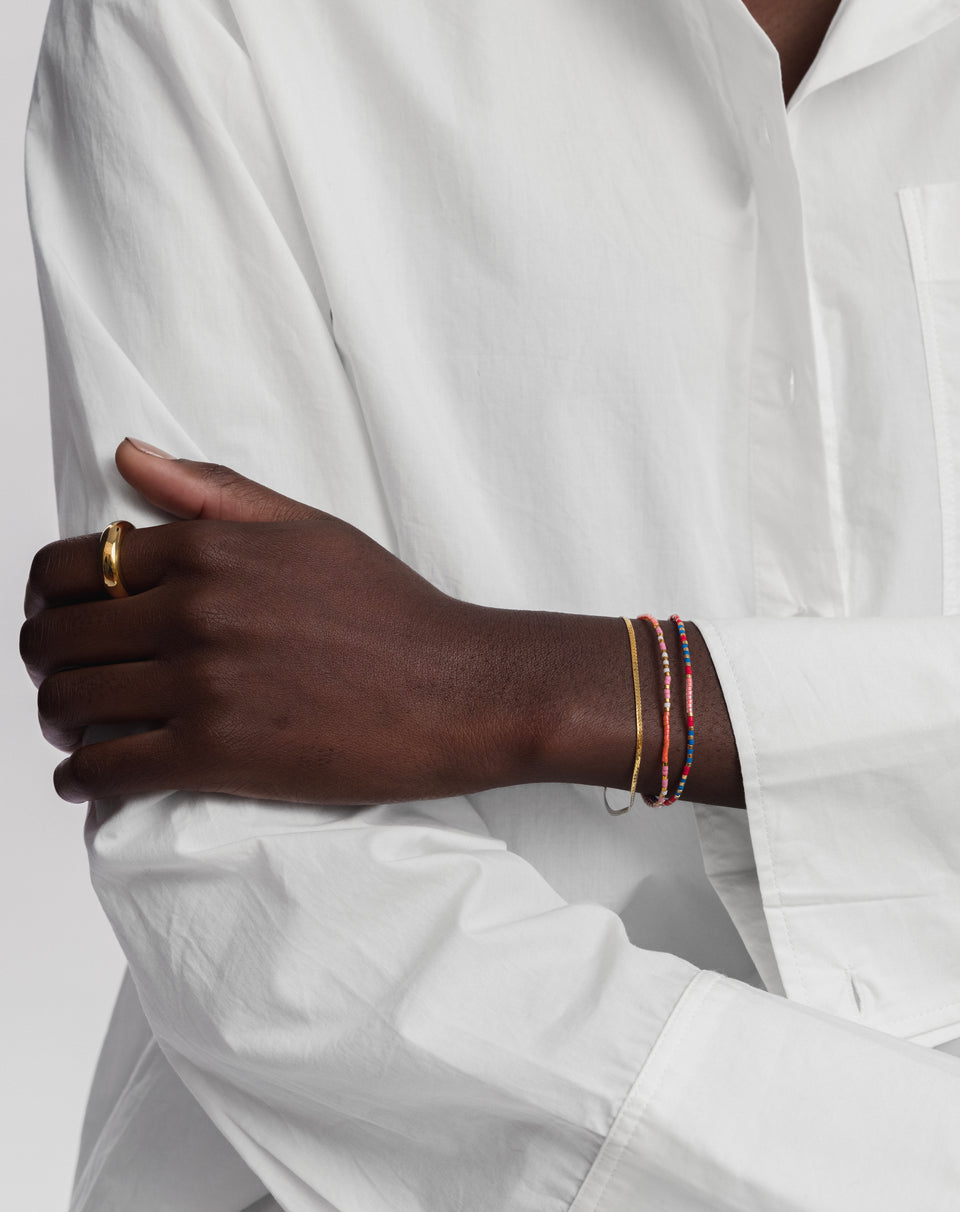 A close-up shot of a model wearing the Herringbone Bracelet in gold, styled with colourful beaded bracelets and a simple gold band ring.