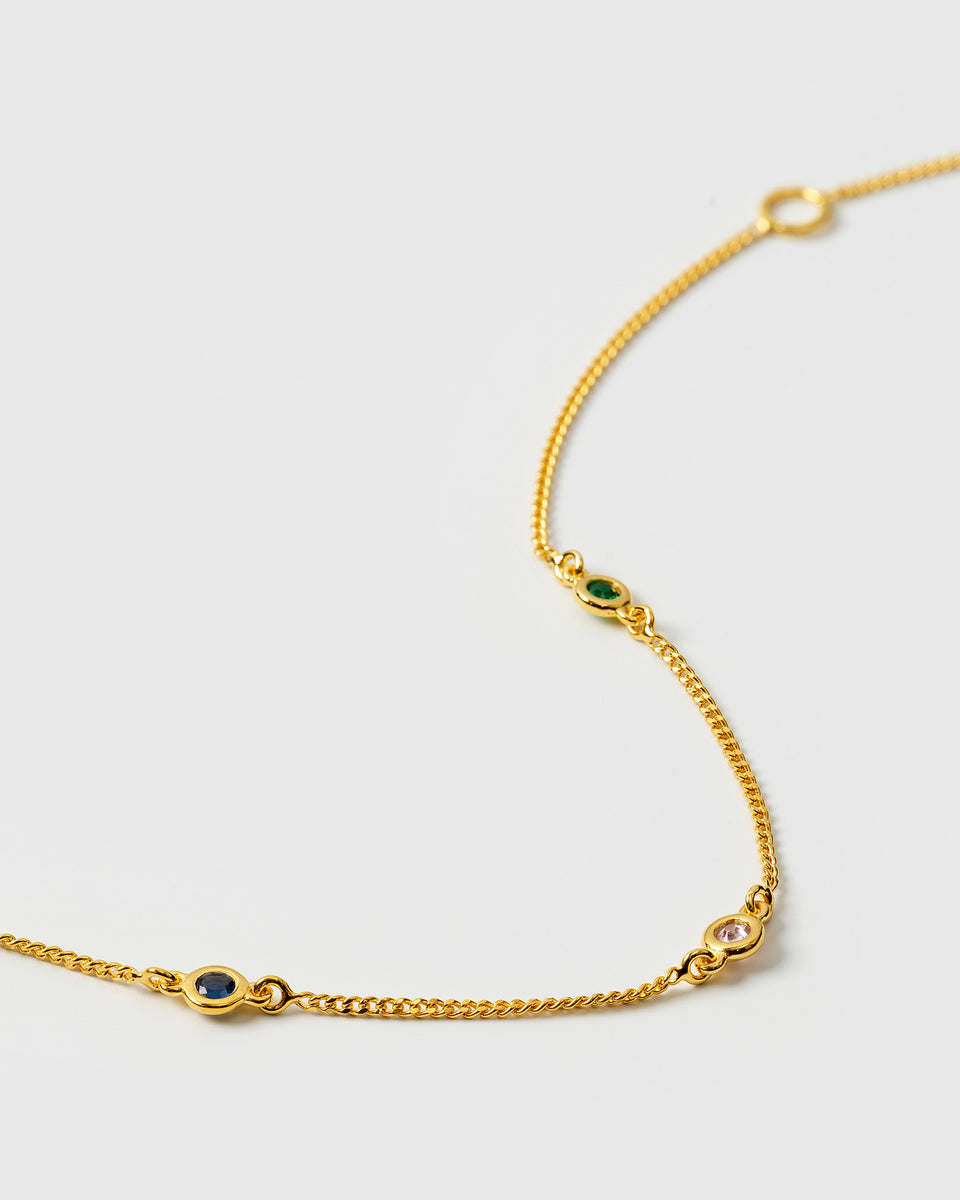 A close-up product shot of the Treasure Shimmer Bracelet in gold, highlighting its fine chain and three faceted zirconias in blue, green, and pink.