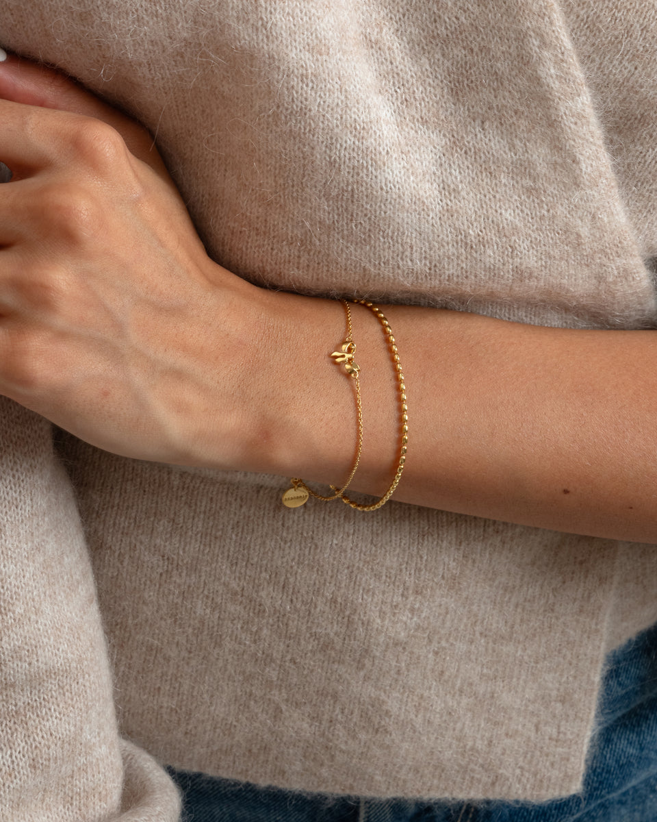 A close-up of a model wearing the Rosie Mini Bracelet in gold, showcasing its delicate bow charm and sparkling, structured chain against her cozy beige sweater.
