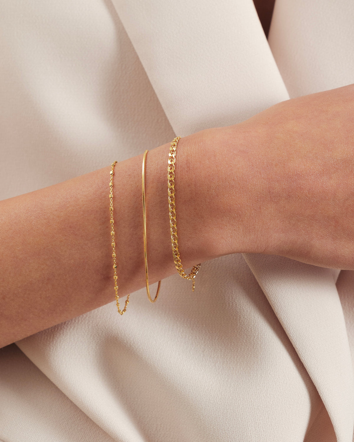 A model's wrist adorned with a stack of three Syster P gold bracelets: the delicate Sheer Station Bracelet, the smooth Sheer Snake Bracelet, and the classic Sheer Curb Bracelet.