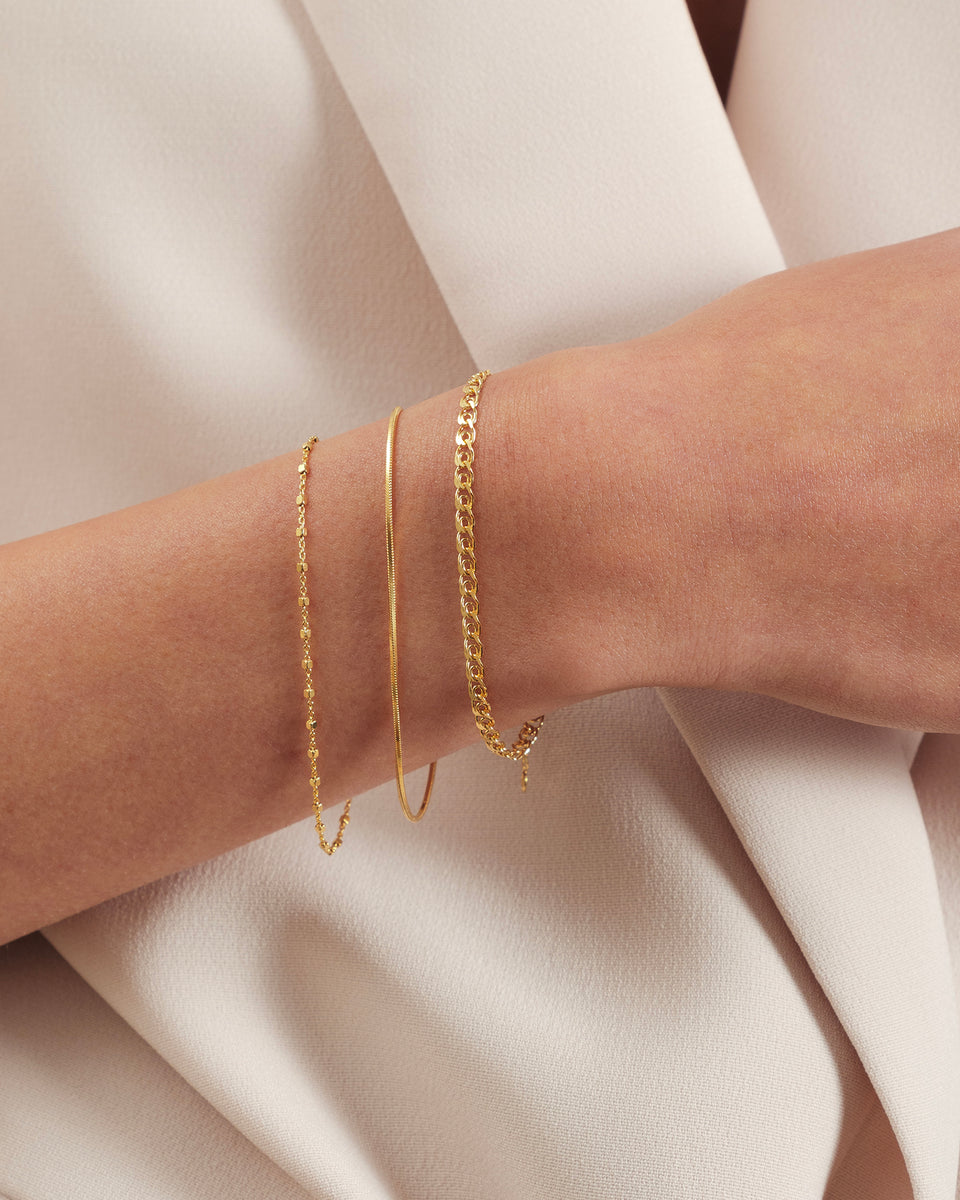 A model's wrist adorned with a stack of three Syster P gold bracelets: the delicate Sheer Station Bracelet, the smooth Sheer Snake Bracelet, and the classic Sheer Curb Bracelet.
