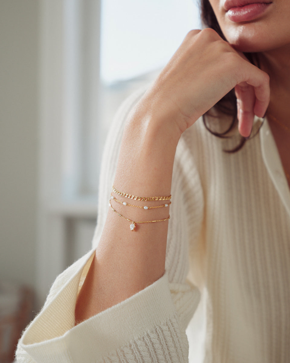 A close-up of a model wearing the Katie Bracelet in gold, featuring a drop-shaped cubic zirconia, layered with a gold curb chain bracelet and a fine gold chain with small pearls.