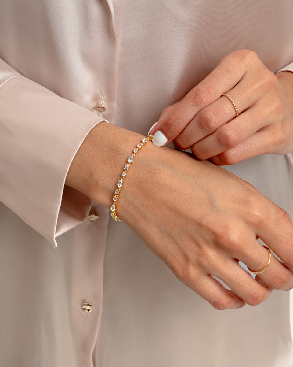 A model wearing the Pam Bracelet in gold on her wrist, which features a pattern of sparkling white topaz stones, styled with two thin gold rings.
