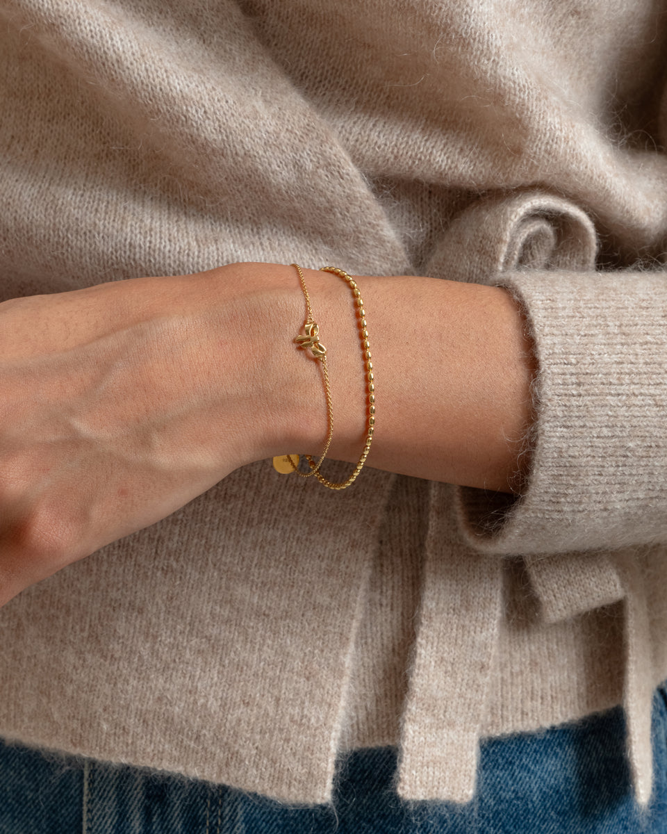 A close-up shot of a model's wrist, showcasing the Lunetta Bracelet in gold, a ball chain design, stacked with a delicate gold chain bracelet featuring a bow charm.