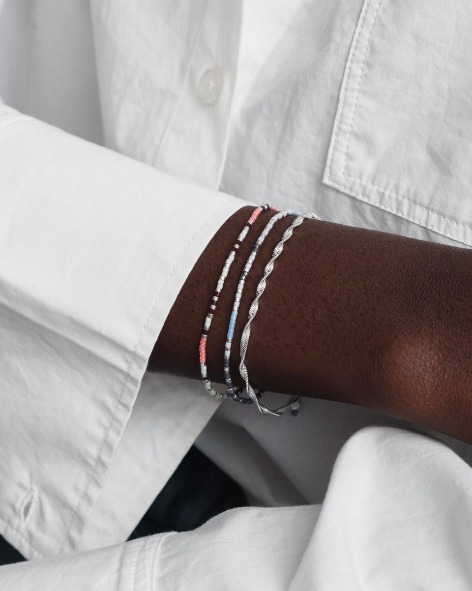 A close-up of a model's wrist wearing the Code Bracelet Silver I Love You, stacked with another blue beaded bracelet and a twisted silver bracelet.