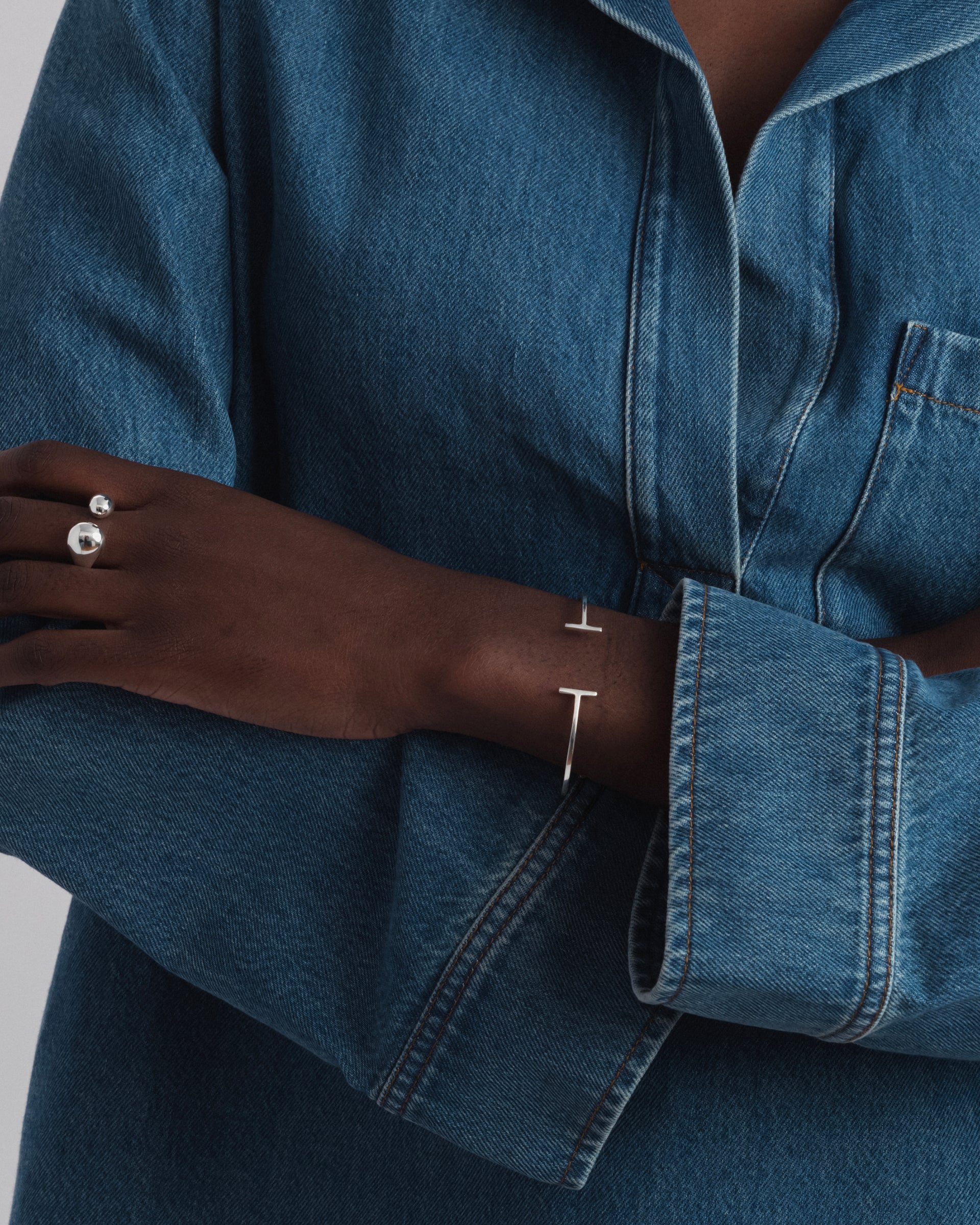 A close-up shot of a model wearing the Strict Bar Bangle in silver on her wrist, along with a silver ring featuring two polished spheres on her finger.