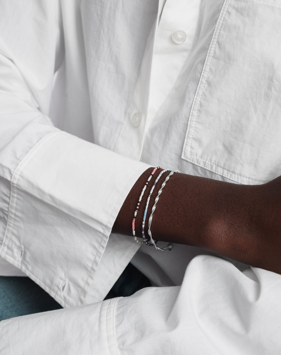 A close-up of a model wearing the Herringbone Twisted Bracelet in silver, stacked with two multi-colored beaded bracelets.