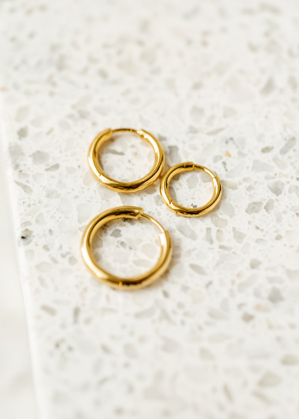 A still life shot of the Beloved Fat Hoops Set in gold, showcasing the three thick hoop earrings in small, medium, and large sizes on a white speckled surface.