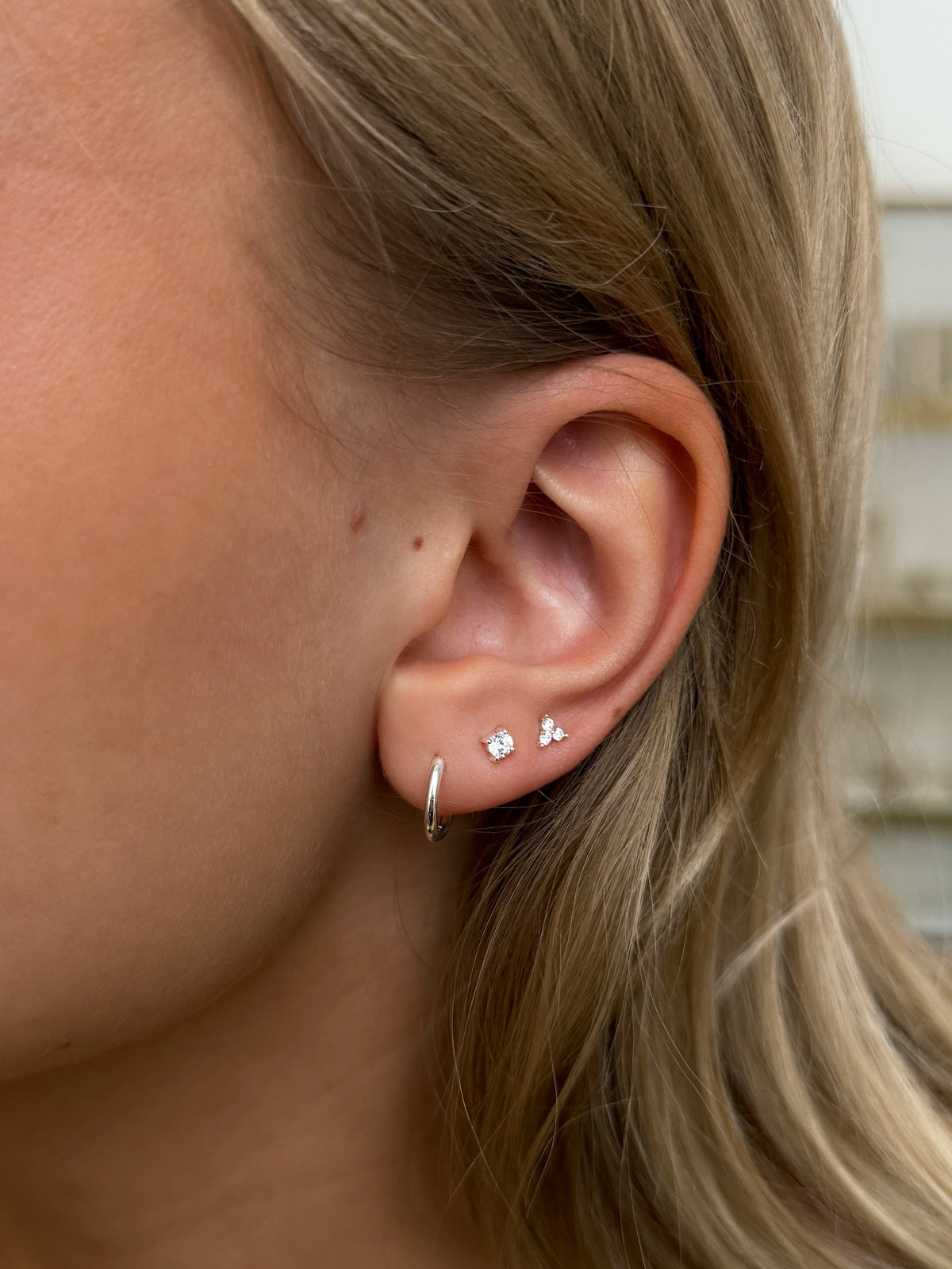A close-up of a stacked silver earrings: a small plain hoop, a single cubic zirconia stud, and a triple cubic zirconia stud.