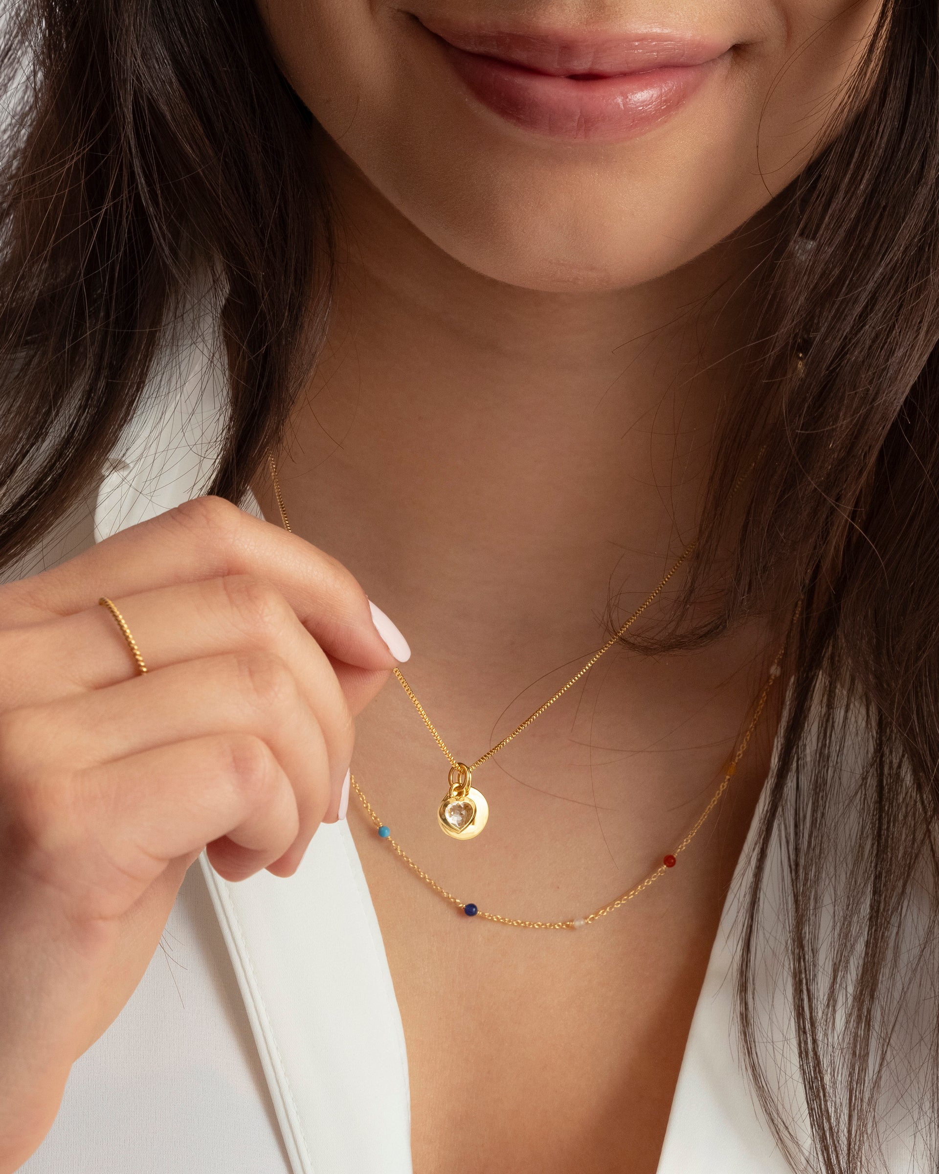 A close-up shot of a model wearing the gold Beloved Heart Pendant with its crystal charm, layered with the gold Santa Monica Necklace adorned with colorful beads, and a delicate twisted gold ring.