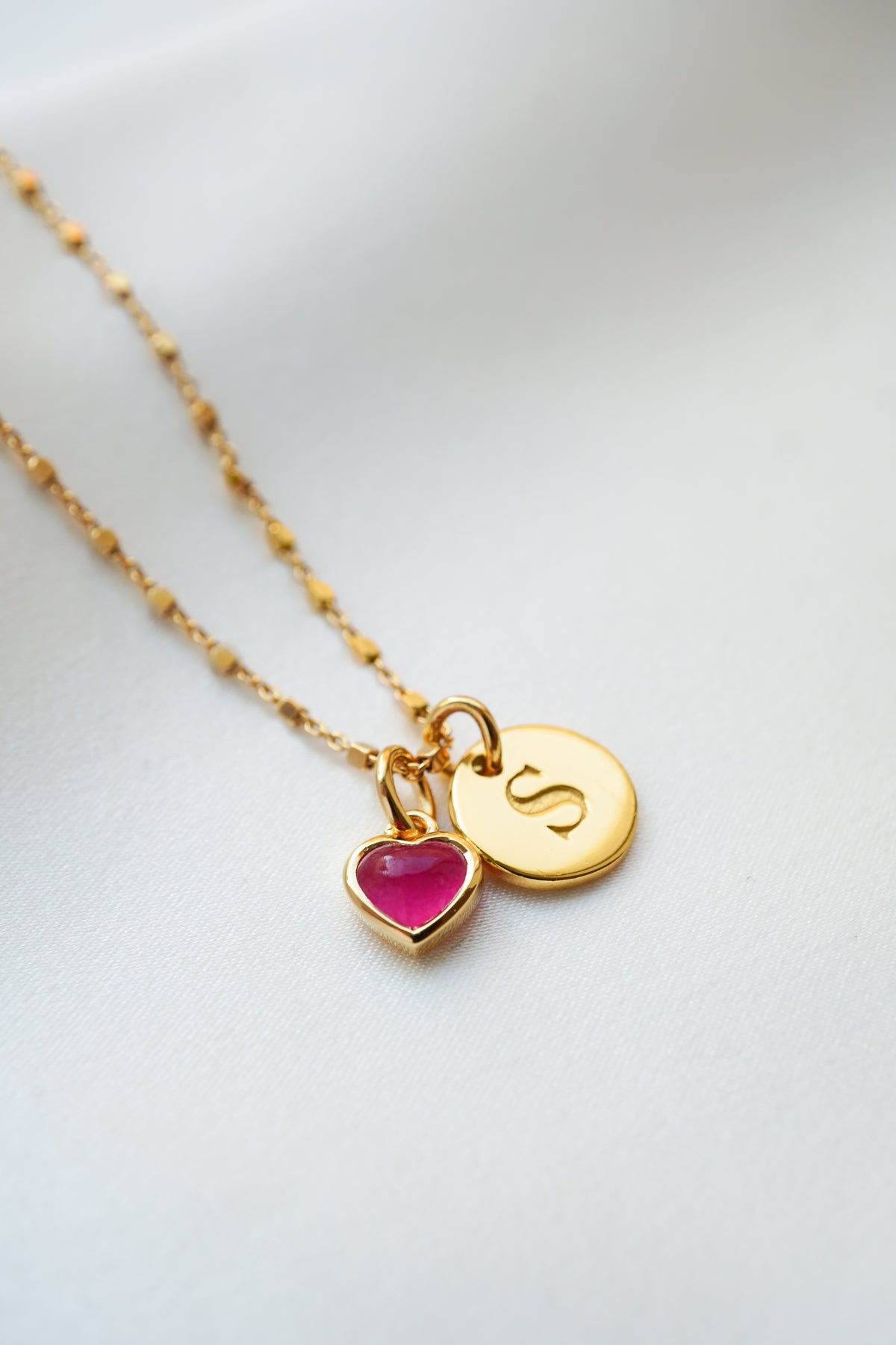 A product shot of the Sheer Station Necklace in gold, styled with the Beloved Heart Pendant in gold with a pink quartz stone, and a gold Beloved Mini Letter 'S' pendant.