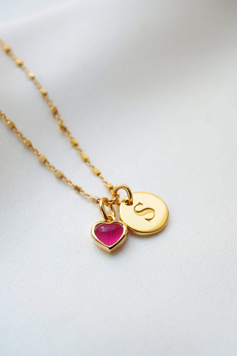 A product shot of the Sheer Station Necklace in gold, styled with the Beloved Heart Pendant in gold with a pink quartz stone, and a gold Beloved Mini Letter 'S' pendant.