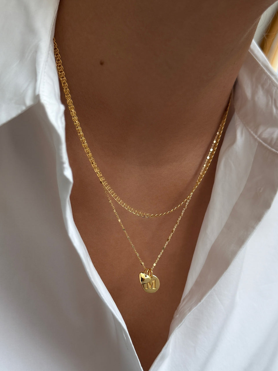 A model wears the gold Sheer Love Necklace, a patterned chain, layered with the longer Sheer Station Necklace featuring a Beloved Heart Pendant and a Beloved Mini Letter 'M' Pendant.