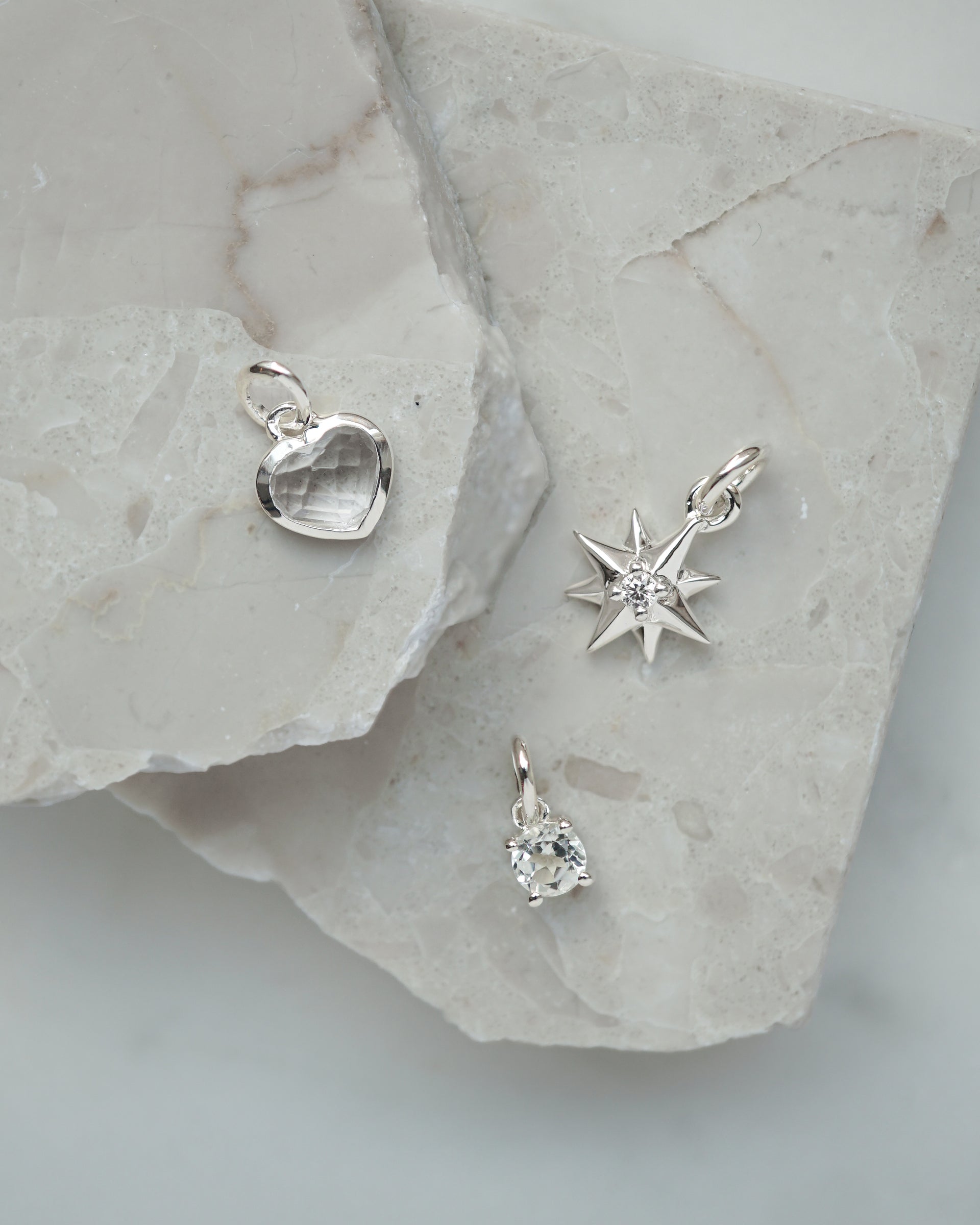 A product shot of three silver pendants from the Syster P Beloved collection: a heart pendant with a clear crystal, a compass star pendant with a central stone, and a small oval crystal pendant.