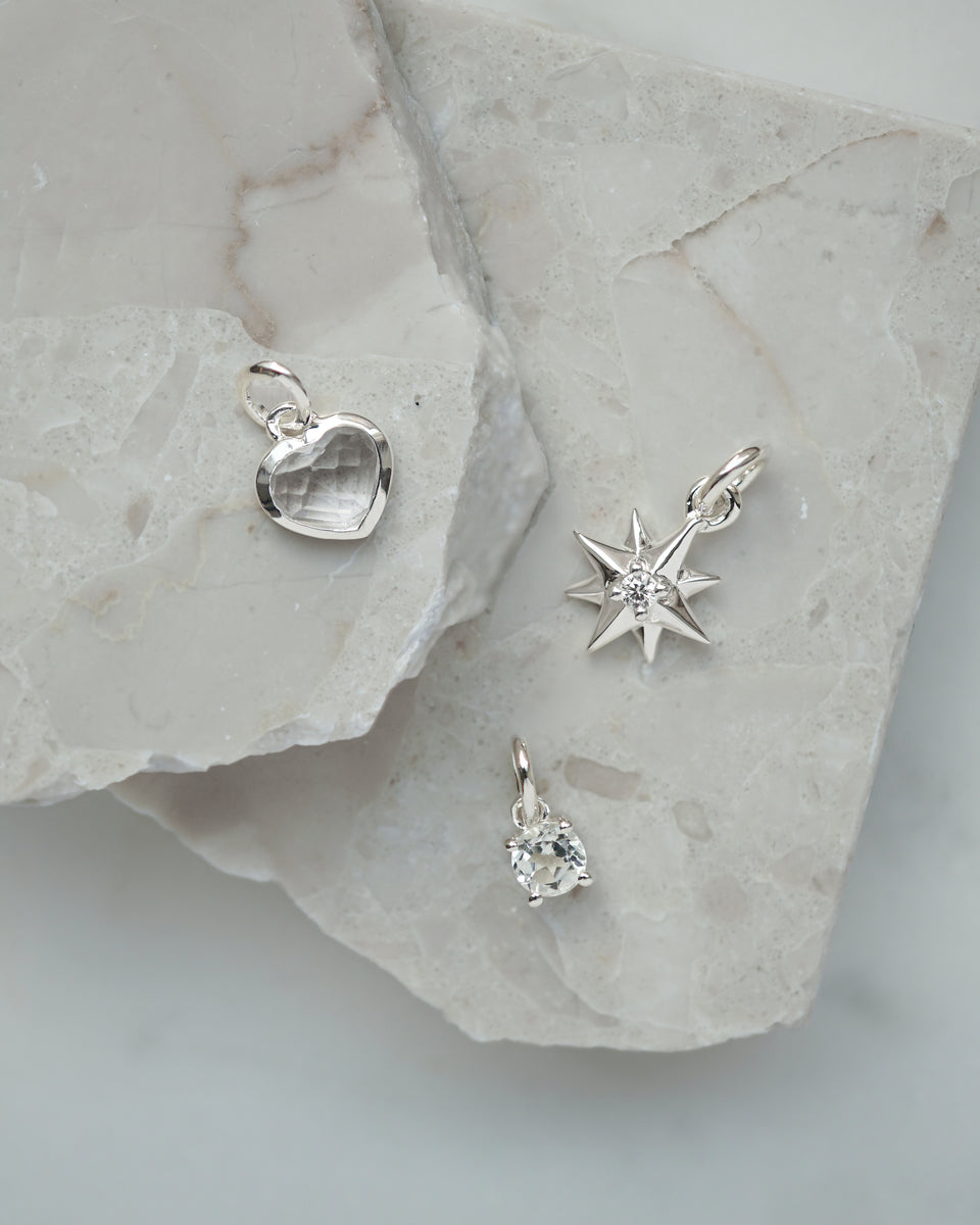 A product shot of three silver pendants from the Syster P Beloved collection: a heart pendant with a clear crystal, a compass star pendant with a central stone, and a small oval crystal pendant.