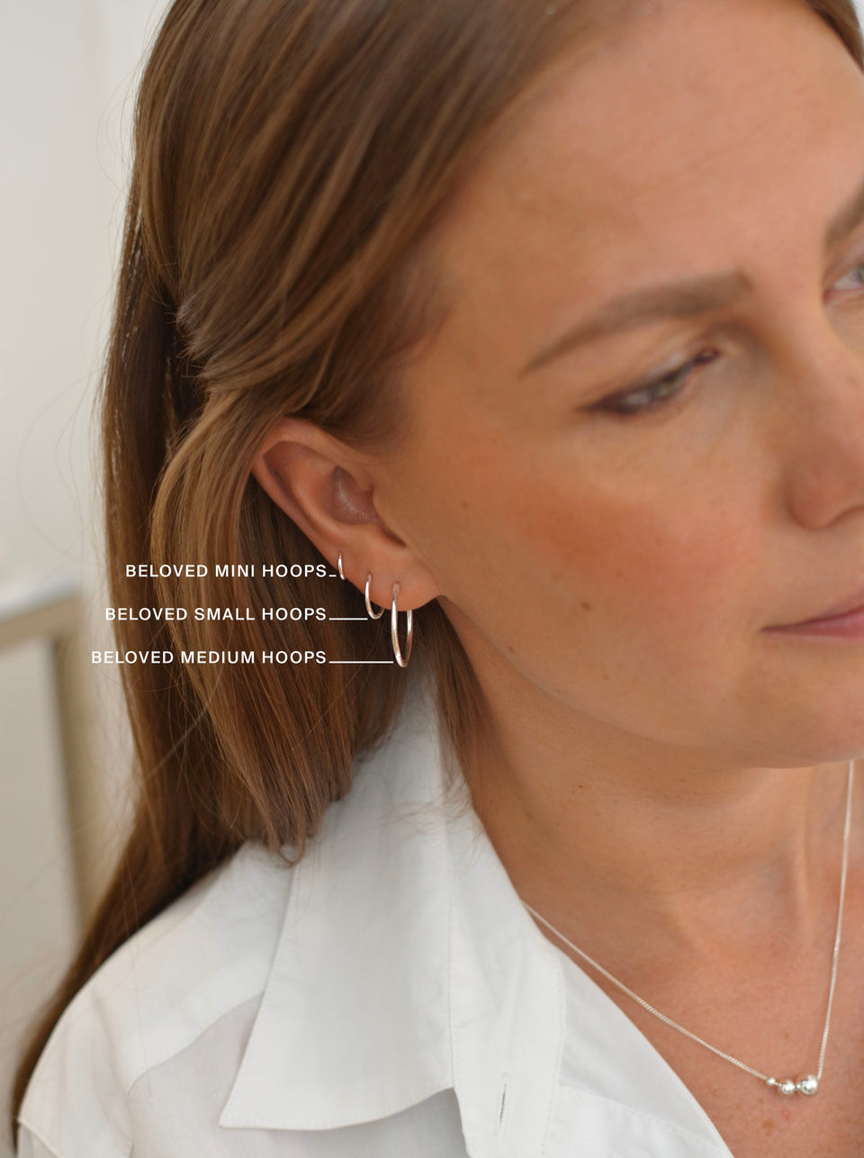 A close-up shot of a model wearing three silver hoop earrings to illustrate the different sizes available: the Beloved Mini, Small, and Medium Hoops. She is also wearing a silver bead necklace.