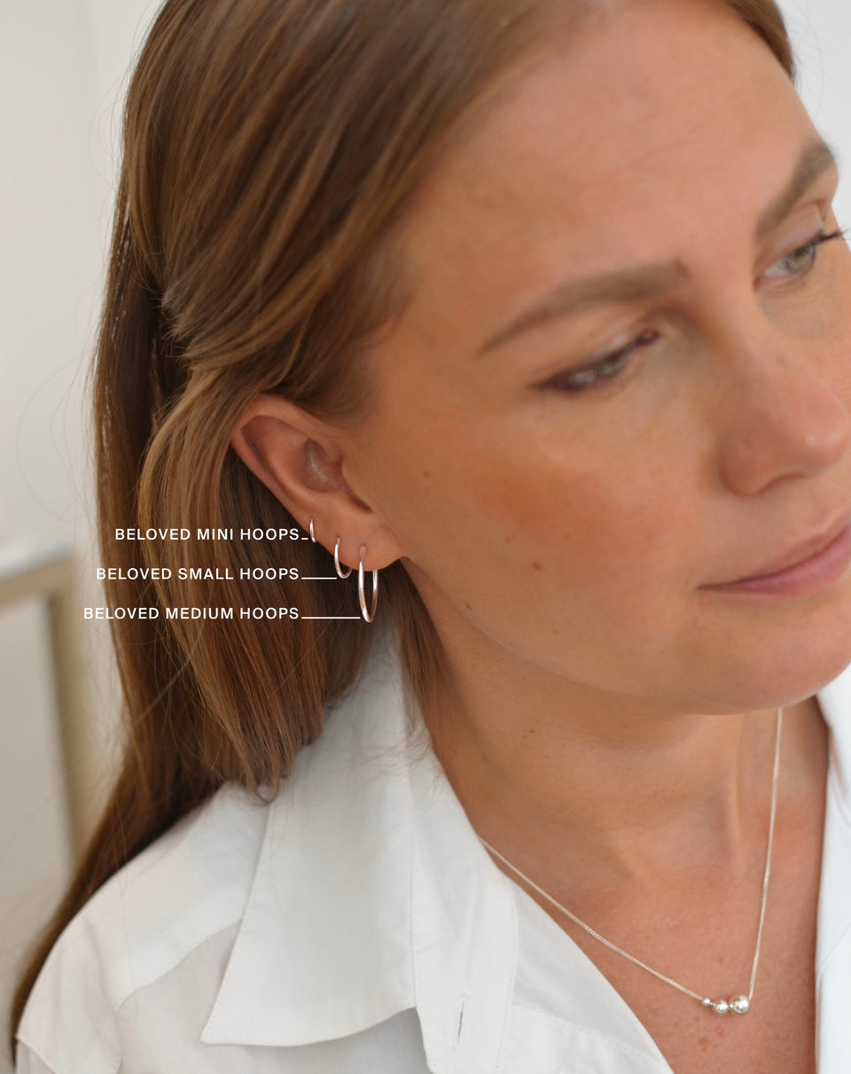 A model wears the Beloved Small Hoops in silver, styled alongside the mini and medium sizes for comparison, and a delicate silver bead necklace.