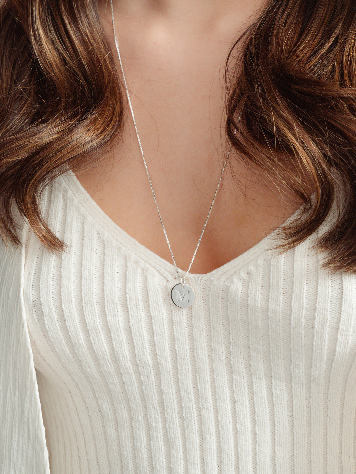 A close-up of a model wearing the Beloved Long Box Chain in silver, paired with a round silver pendant engraved with the letter M.