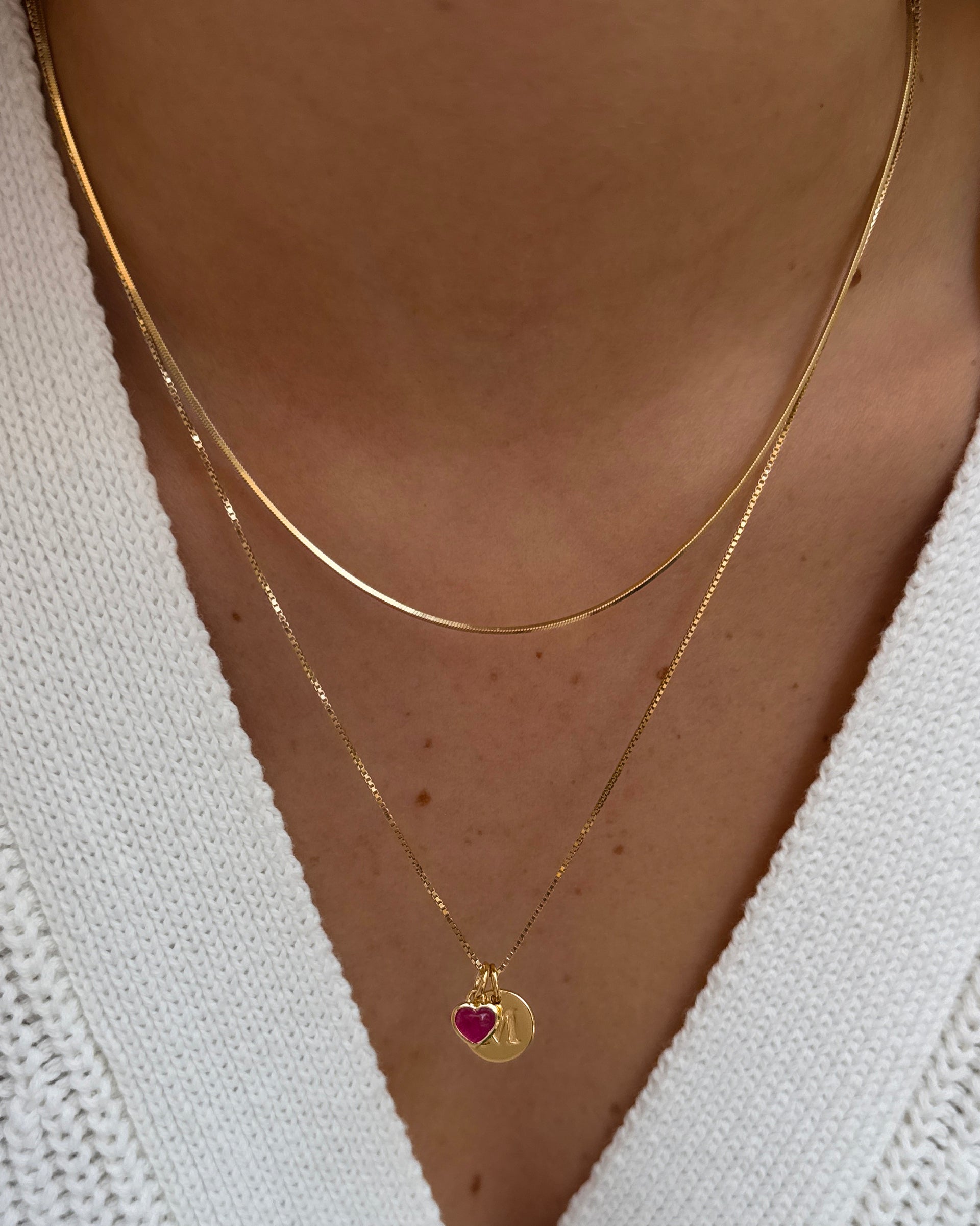 A close-up shot of a model wearing a layered necklace combination, featuring the gold Sheer Mirror Necklace and a gold Beloved Box Chain with a Beloved Heart Pendant in pink quartz and a mini letter pendant.