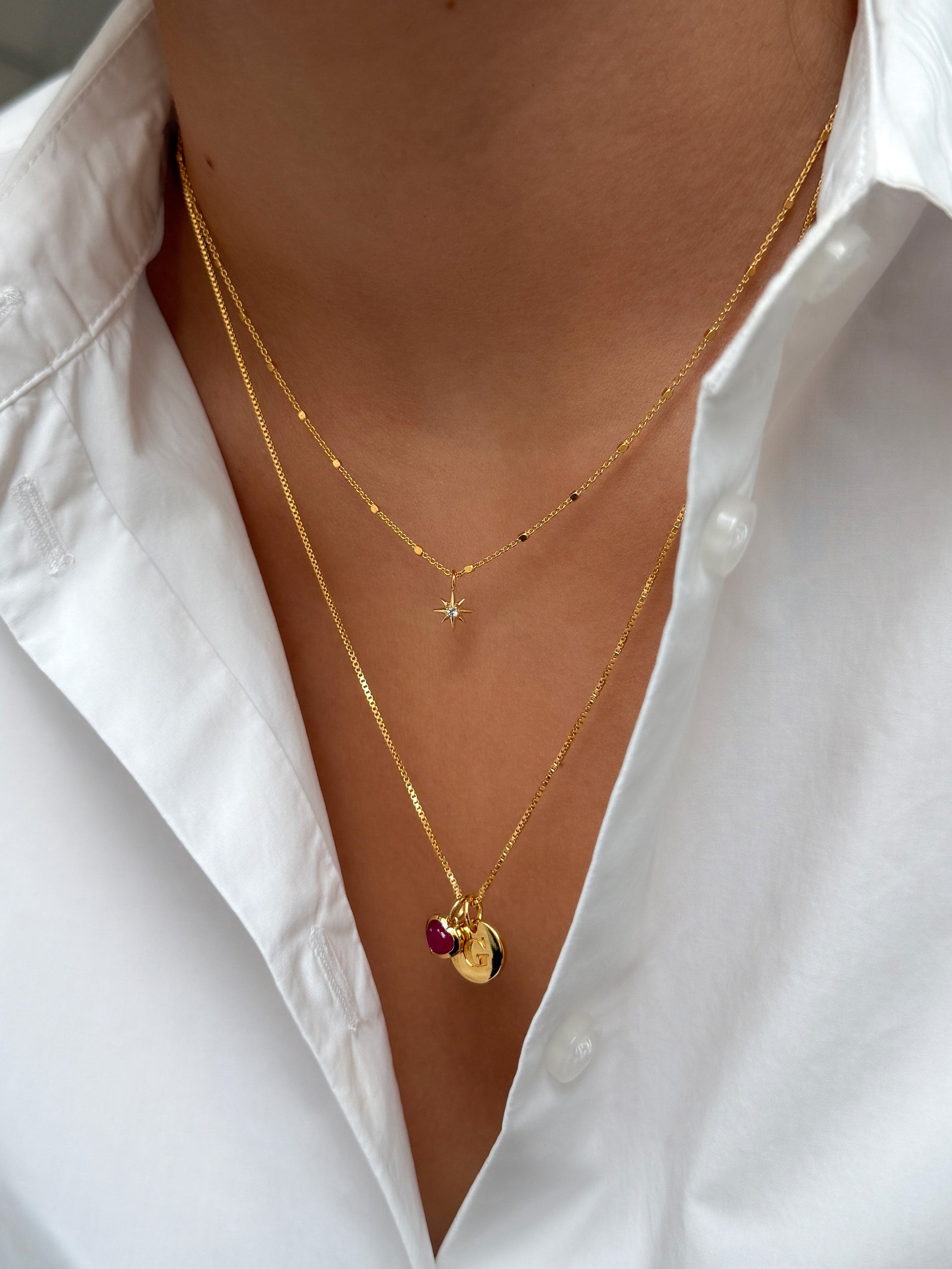 A close-up shot of a model wearing two layered gold necklaces: the shorter North Star Necklace with a star pendant, and a longer box chain necklace with a pink heart and a letter charm.