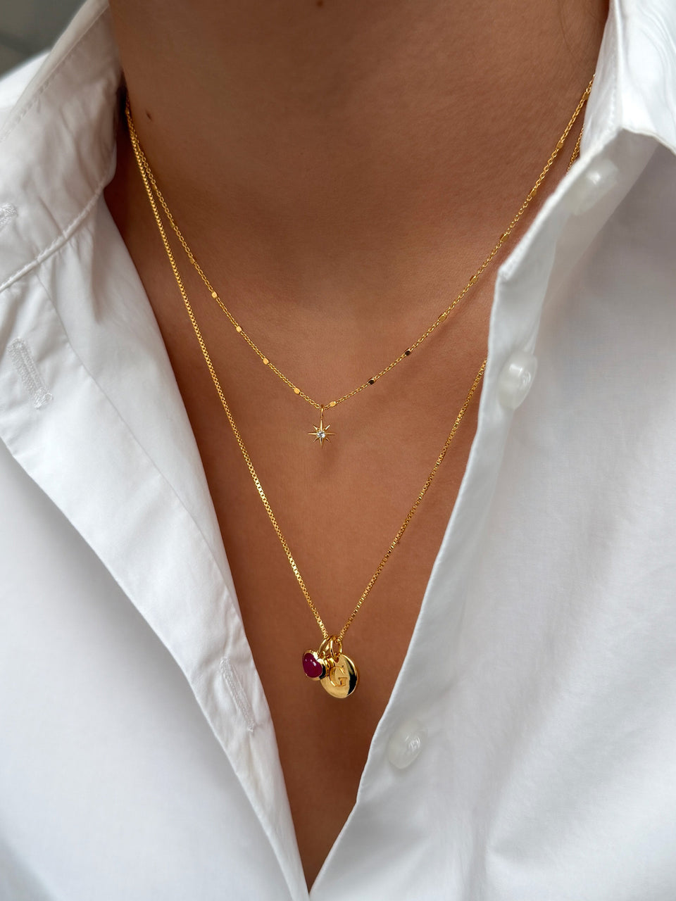 A close-up shot of a model wearing two layered gold necklaces: the shorter North Star Necklace with a star pendant, and a longer box chain necklace with a pink heart and a letter charm.