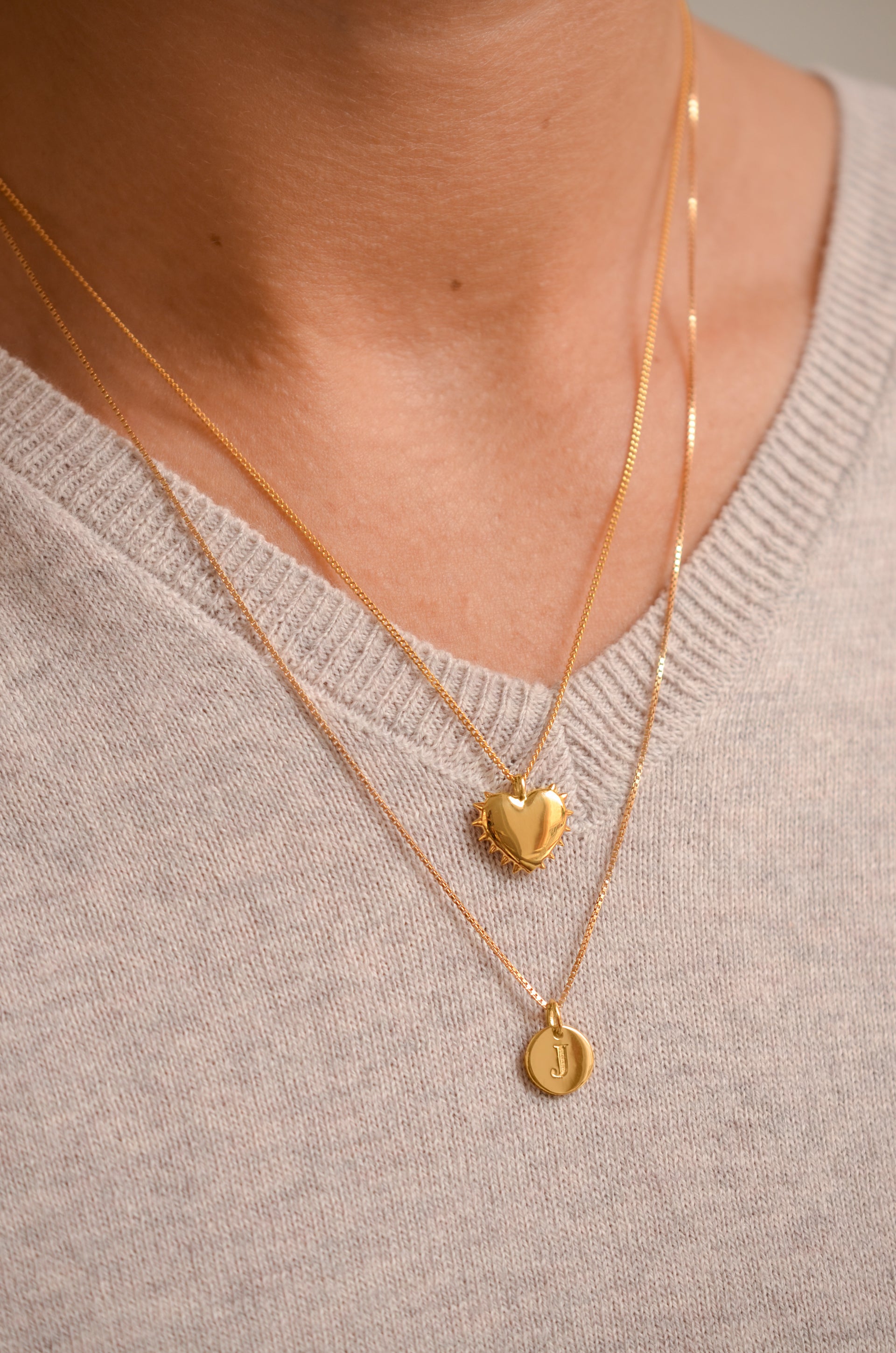 A close-up shot of a model wearing two layered Syster P gold necklaces against a fine-knit grey sweater. The top necklace is the True Love Spike Heart Necklace, featuring a heart pendant with a spiky edge, layered with a longer Beloved Medium Box Chain featuring a Beloved Mini Letter 'J' pendant.