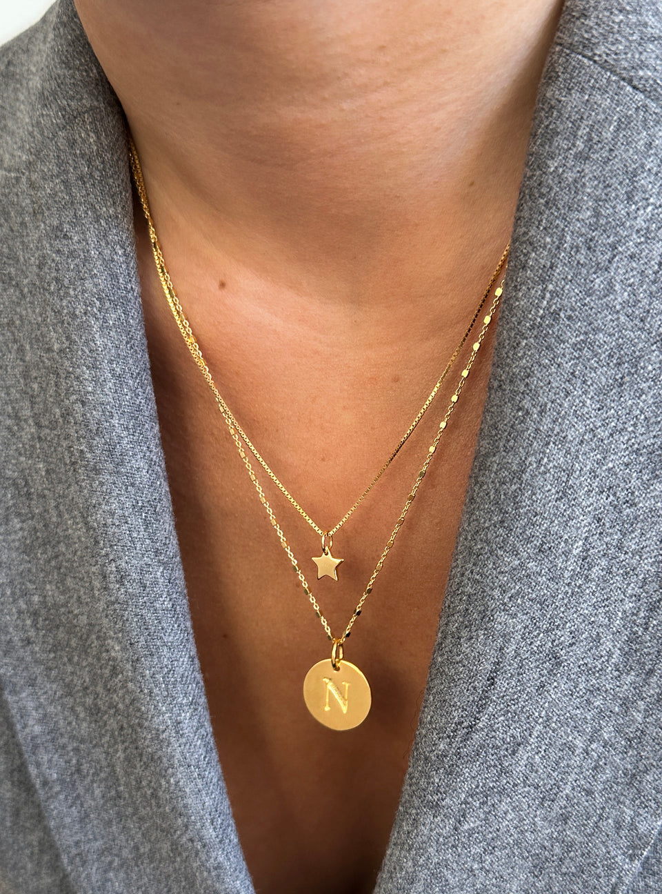 A close-up of a model wearing two layered gold necklaces. The shorter necklace features a small star pendant on a box chain, and the longer one has a round pendant with the letter 'N' on a station chain.
