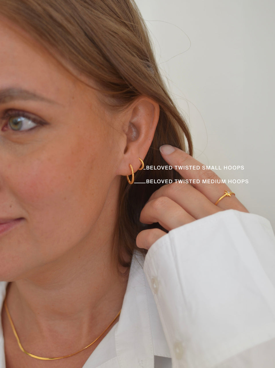 A close-up shot of a model wearing both the Beloved Twisted Small and Medium Hoops in gold to show the size difference. The look is completed with a gold-colored snake chain necklace and a knot ring.