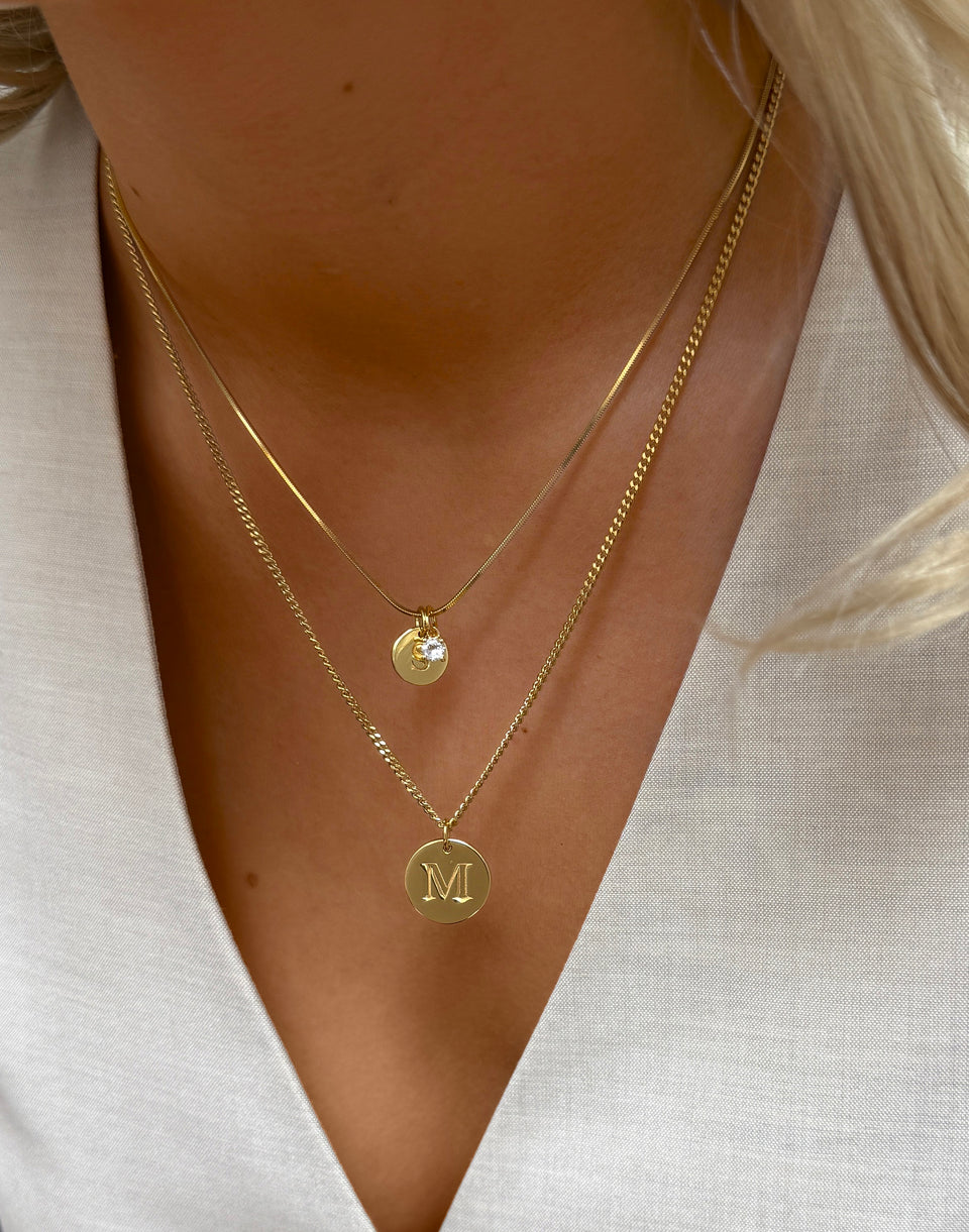 A close-up shot of a model's neck, showcasing two layered gold necklaces. The shorter necklace has a delicate snake chain with two charms: a small gold disc with the letter 'S' and a sparkling white topaz pendant. The longer necklace features a curb chain with a larger gold disc pendant engraved with the letter 'M'.