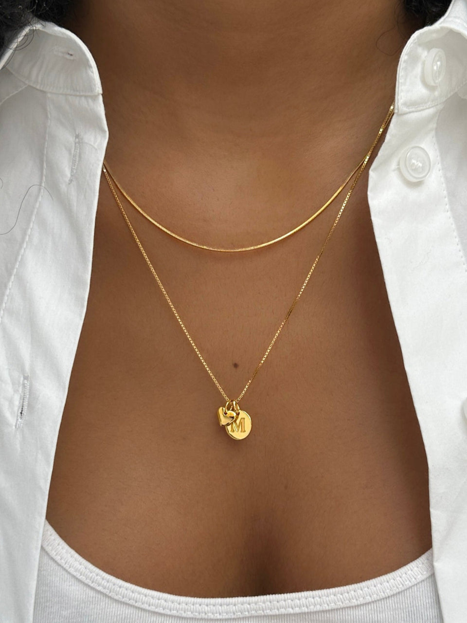 A close-up shot of a model wearing two layered gold necklaces: the Sheer Mirror Necklace and the Beloved Medium Box Chain, which is personalized with a Beloved Heart Pendant and a Beloved Mini Letter 'M' pendant.