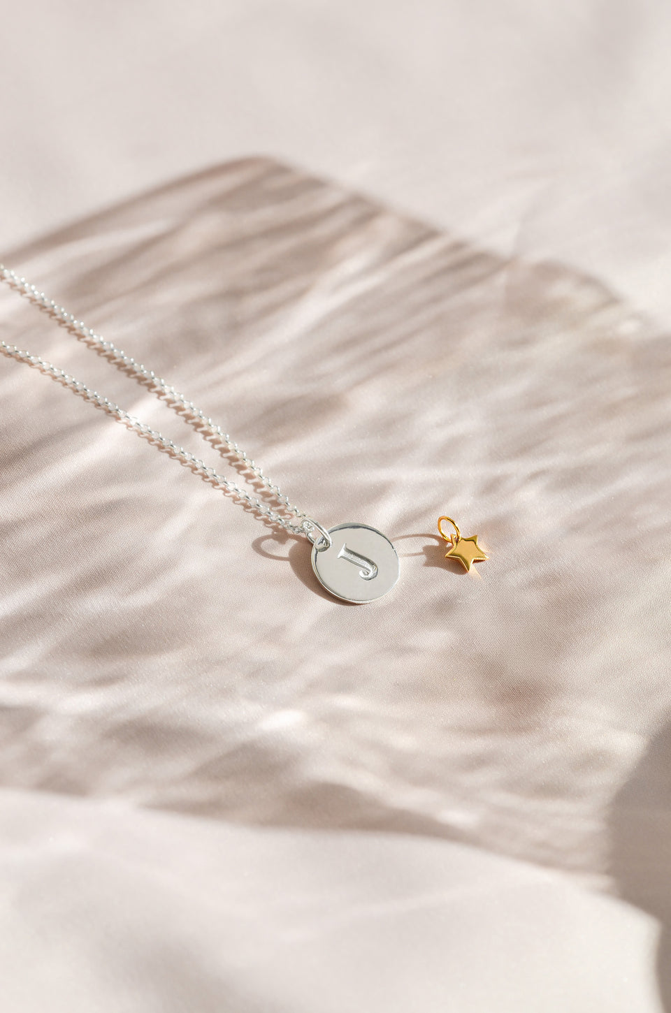 A product shot of the Beloved Star Pendant in gold, styled alongside a silver necklace with a round pendant engraved with the letter 'J'.