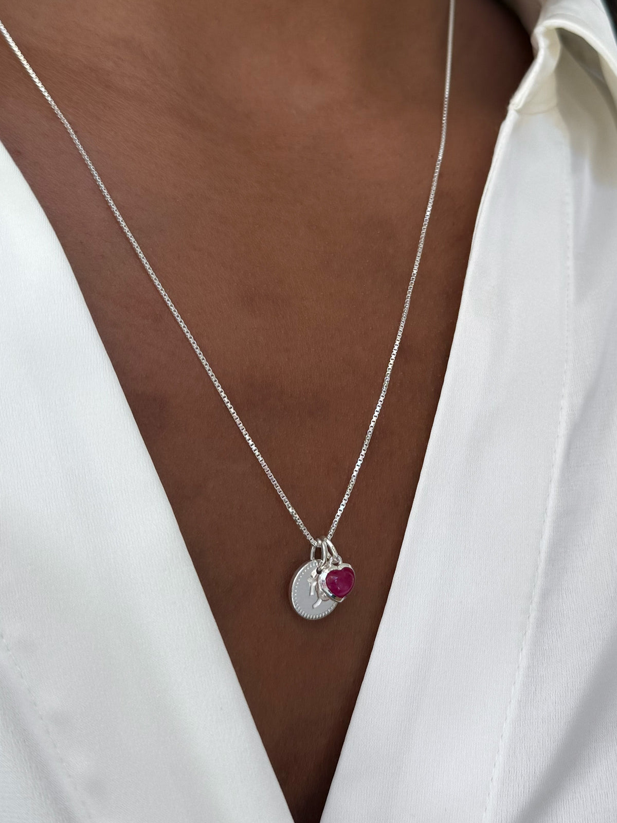 A close-up on a model wearing a silver box chain necklace, styled with a round silver Capricorn zodiac pendant and a small silver heart pendant with a pink stone.