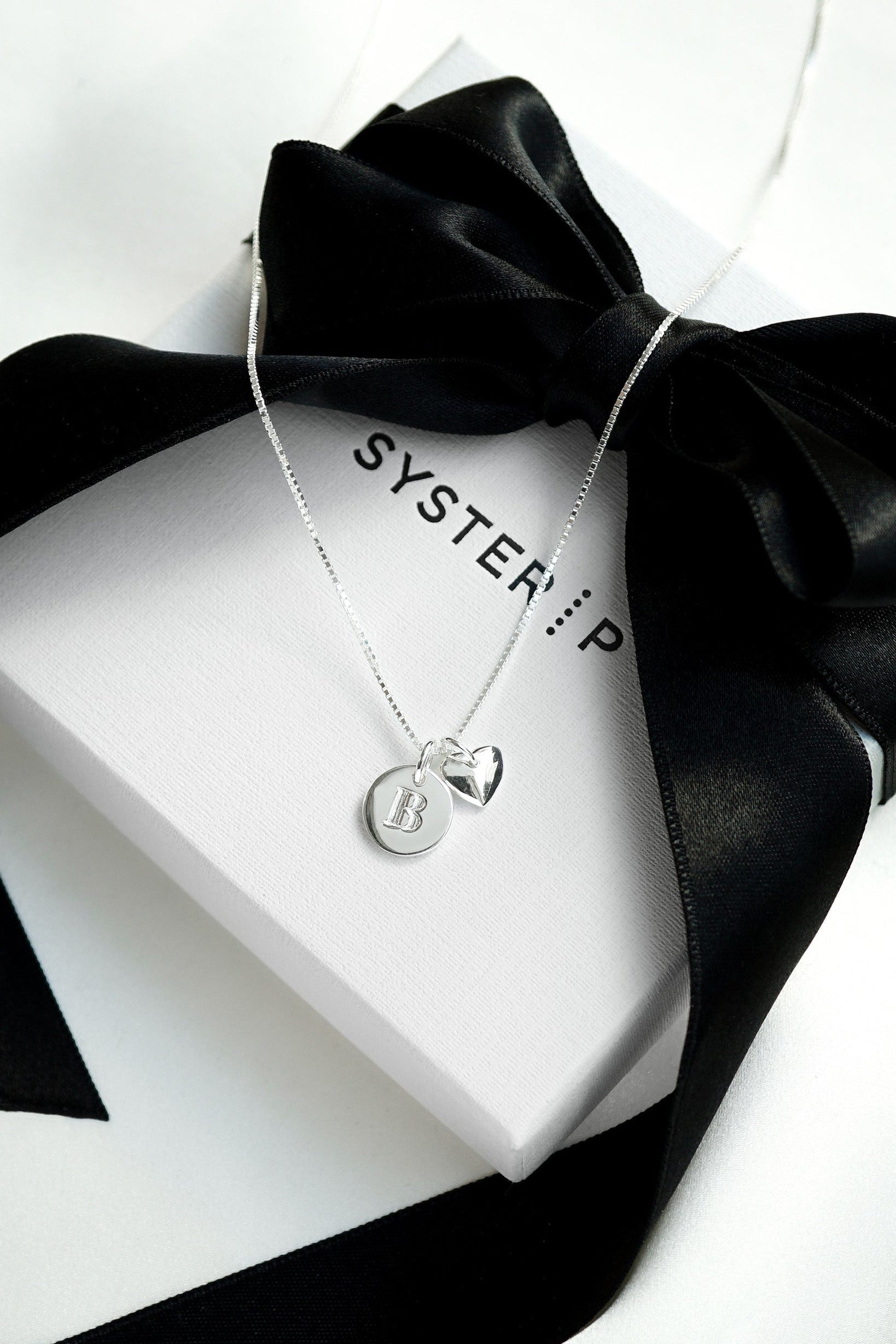 A product shot of a silver Syster P Beloved necklace with a medium box chain, featuring a mini letter 'B' pendant and a heart pendant, resting on a white gift box with a black ribbon.