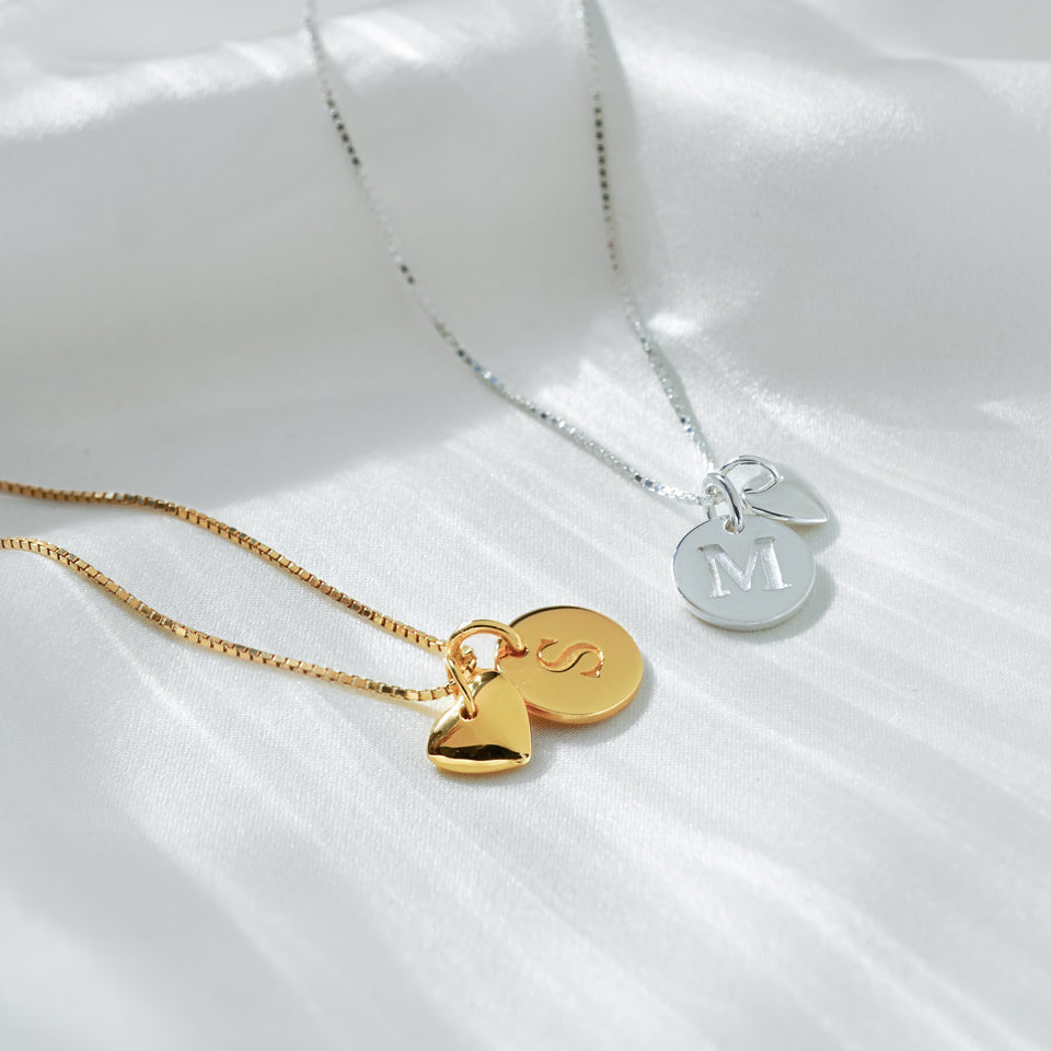 A product shot of two Beloved necklaces, one in gold with a heart pendant and a mini 'S' letter pendant, and one in silver with a heart pendant and a mini 'M' letter pendant, both on box chains.