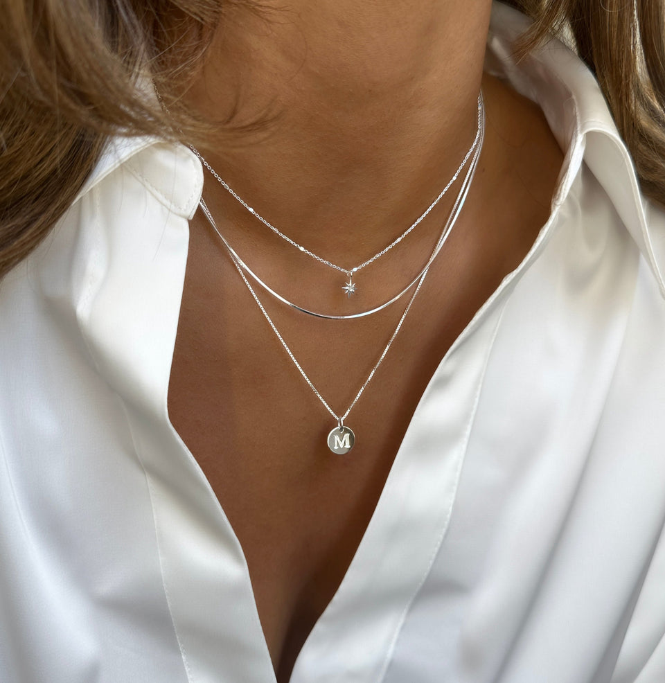 A model wearing a layered look with three Syster P silver necklaces: a fine chain with a North Star pendant, a flat Sheer Mirror chain, and the Beloved Medium Box Chain with a round letter 'M' pendant.