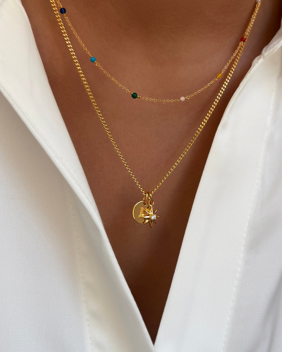 A close-up of a model wearing two layered gold necklaces against a white shirt. The longer necklace features the Beloved Compass Star Pendant alongside a mini letter pendant, while the shorter one is adorned with small colorful beads.