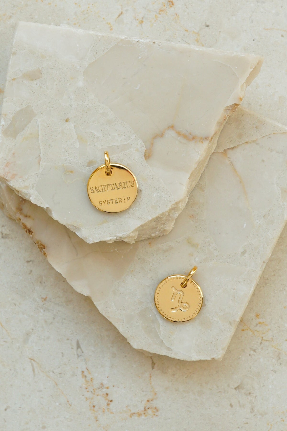 A product shot of two gold Beloved Zodie Pendants on a marble surface, one showing the front with the engraved Capricorn symbol and the other showing the back with the engraved word 'Sagittarius'.
